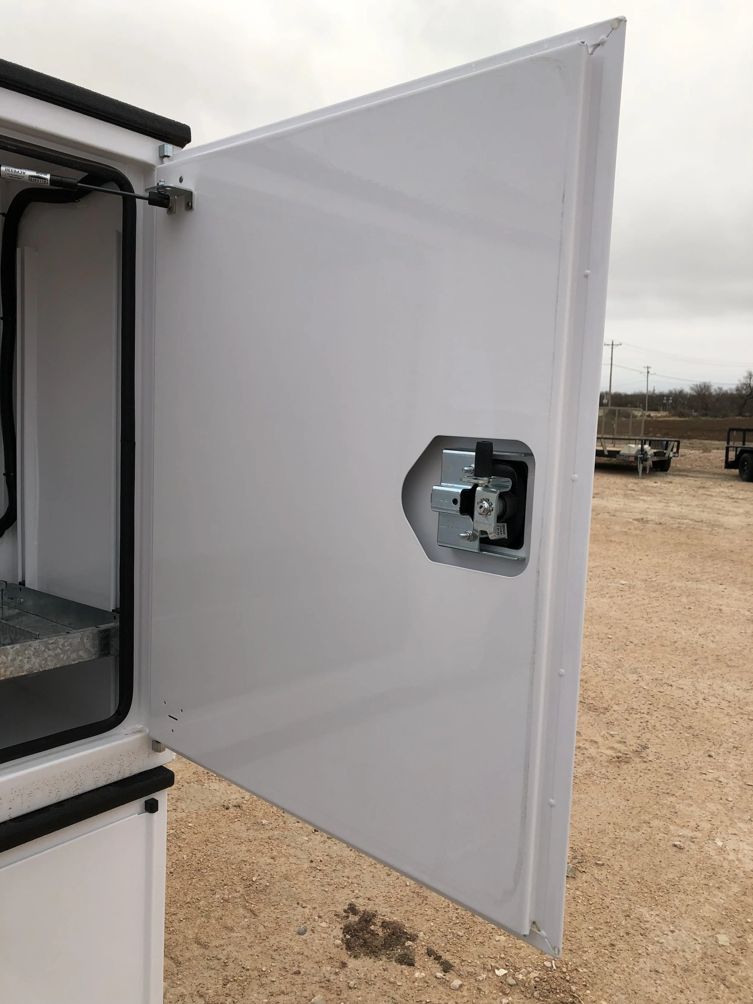 Open white metal door with latch, on an outdoor utility or service truck, dirt ground and overcast sky in background.