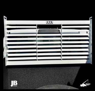 A large, white metal gate with horizontal slats, mounted on a black surface, with a handle in the center. The gate has small logos and initials on the bottom left and right corners.