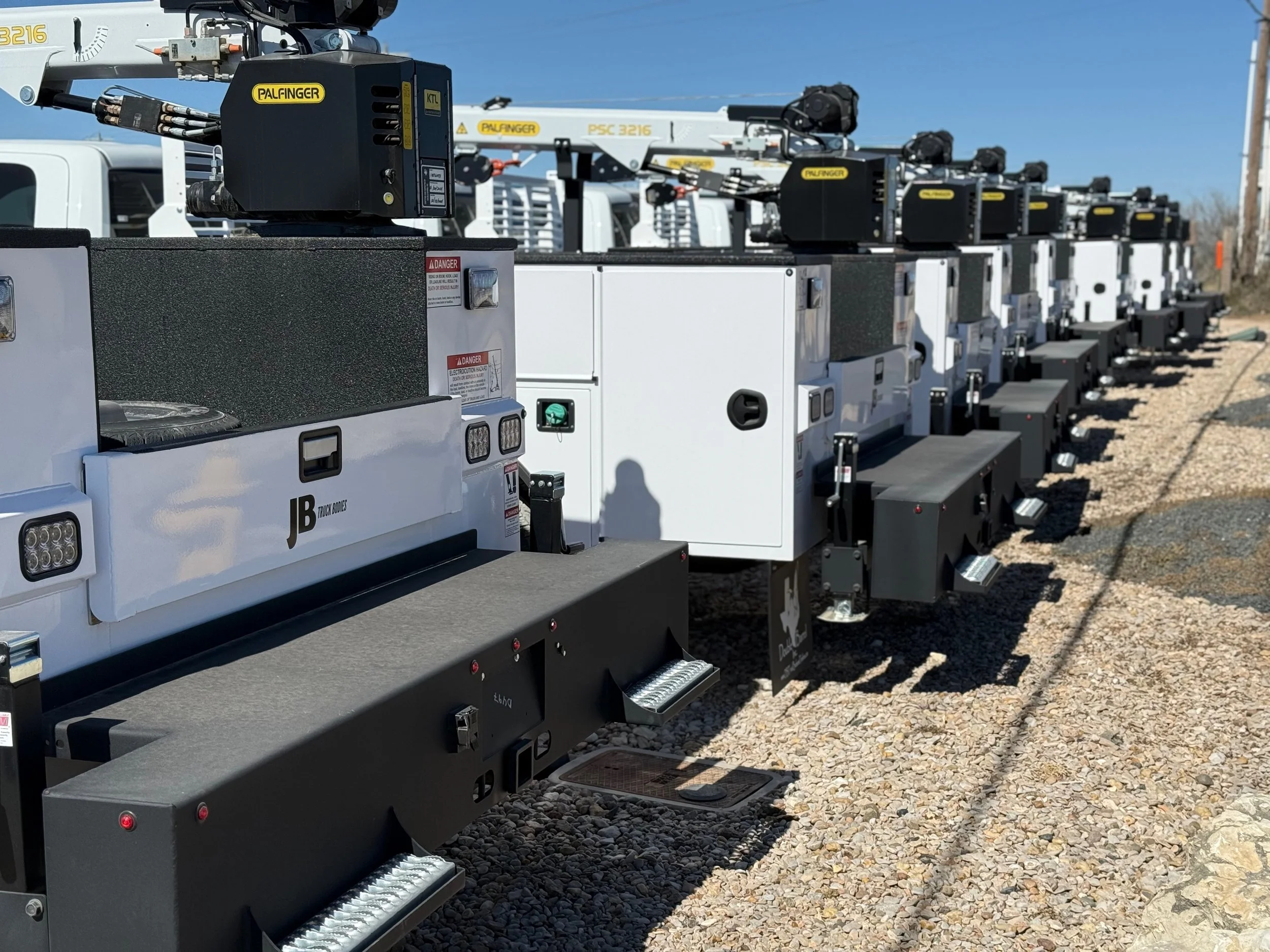 Row of crane truck bodies and lifting equipment parked outdoors on gravel under a clear blue sky.