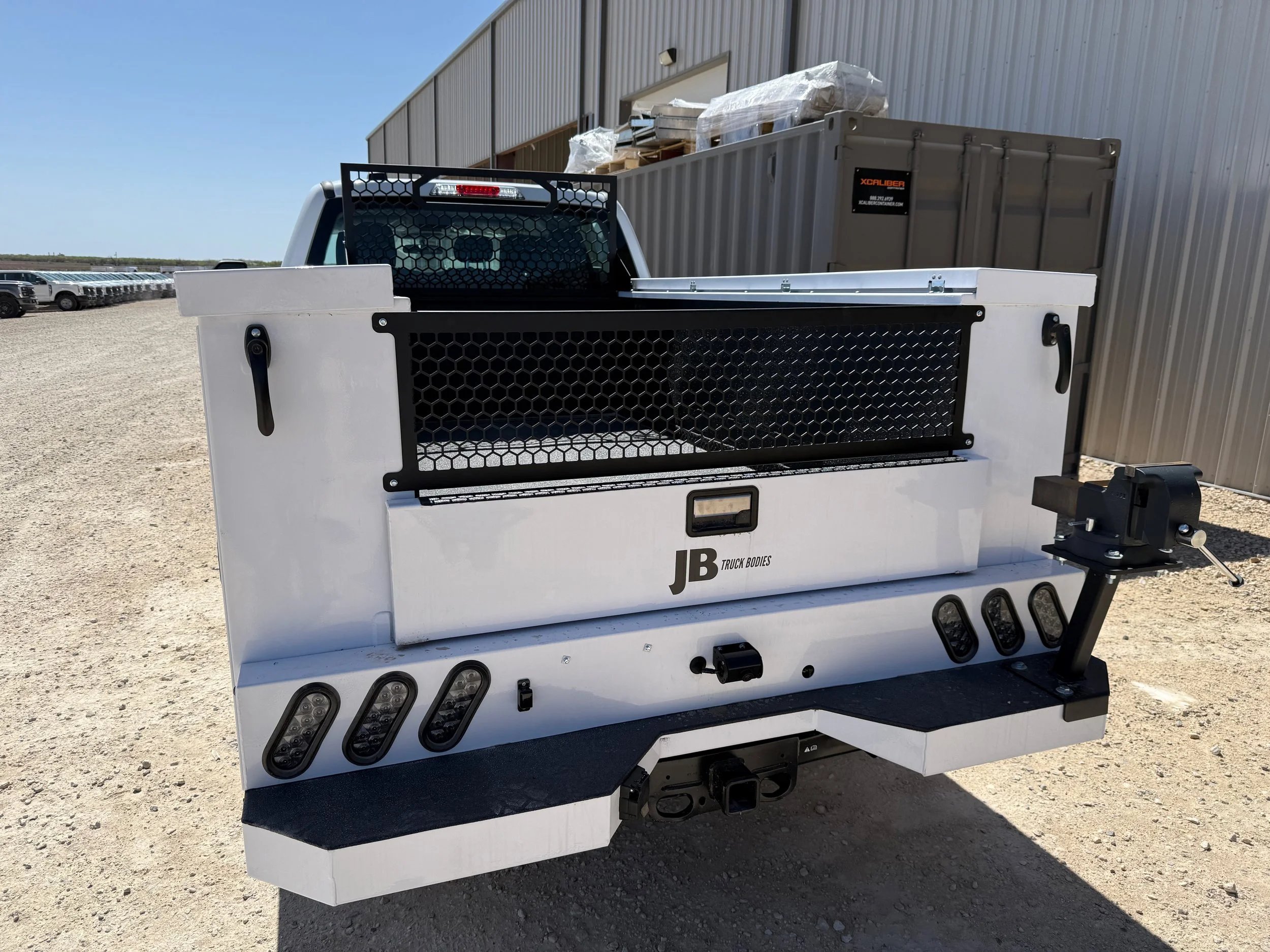 The rear of a white utility truck bed with various compartments and a mounted work device, parked outdoors near a metal building.