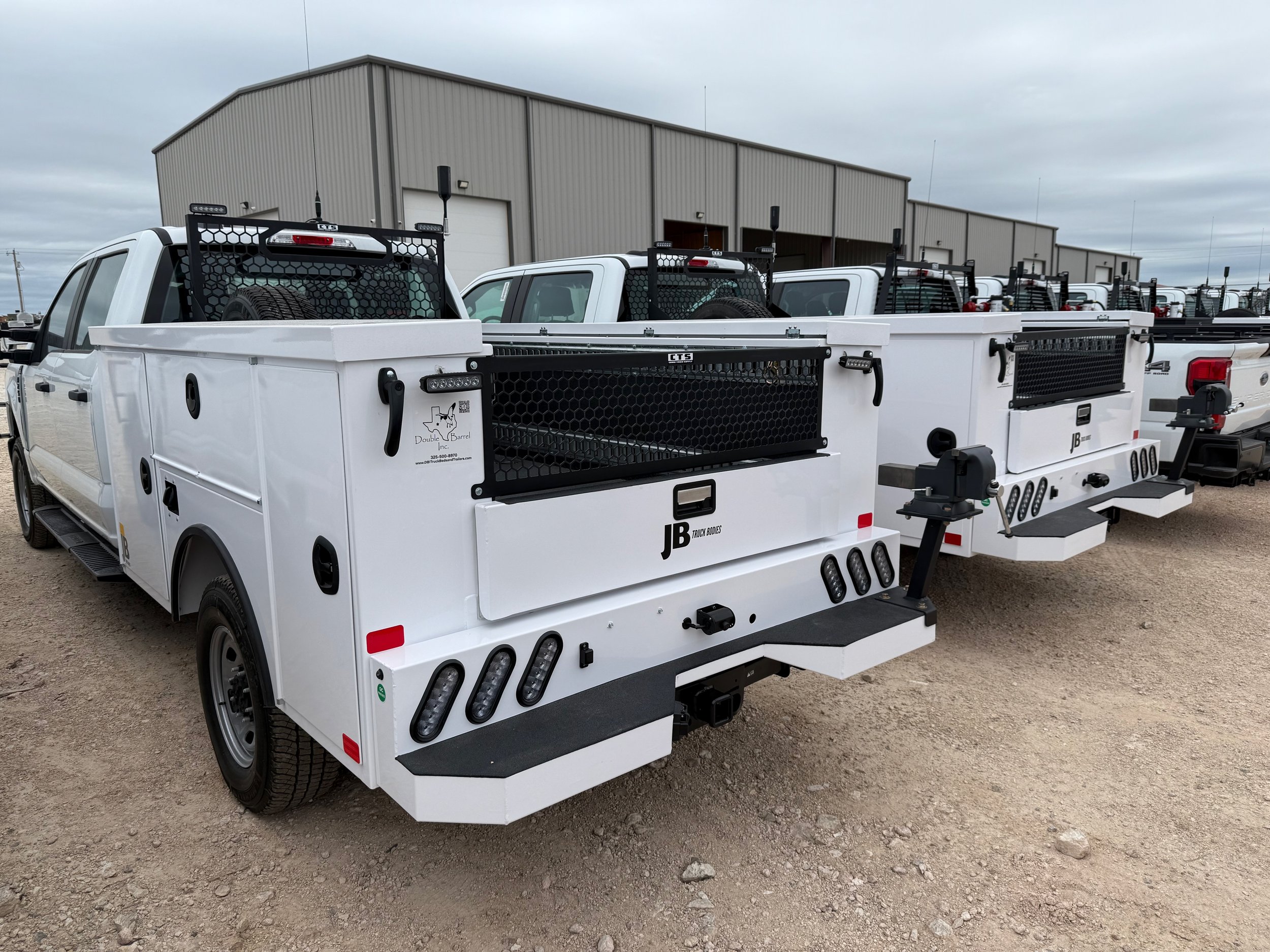 Two white utility trucks with tool storage compartments and racks, parked outdoors on a gravel lot with a large metal building in the background.