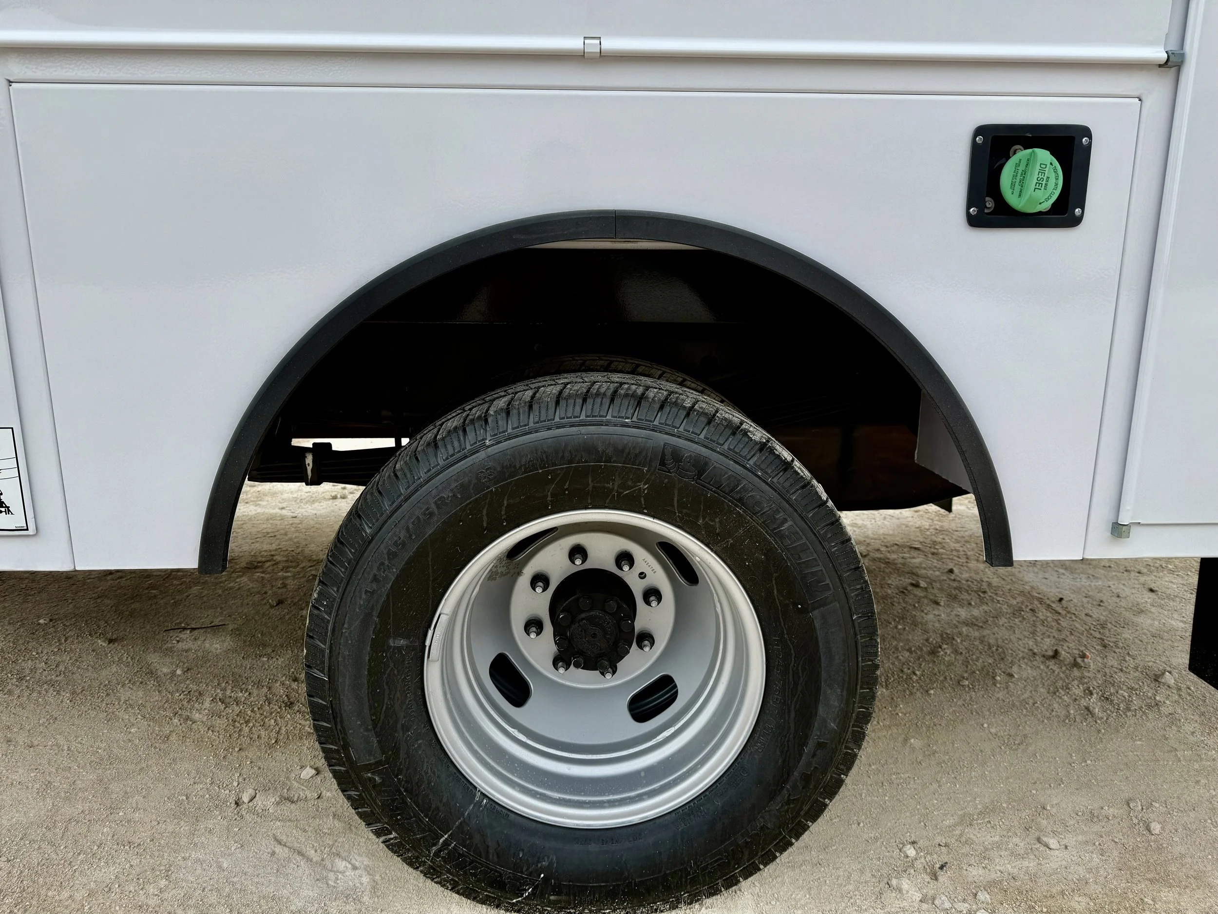 Close-up of a vehicle wheel on an off-road surface, showing the tire, rim, and part of the white vehicle body with a black wheel well and a green circular valve cap.