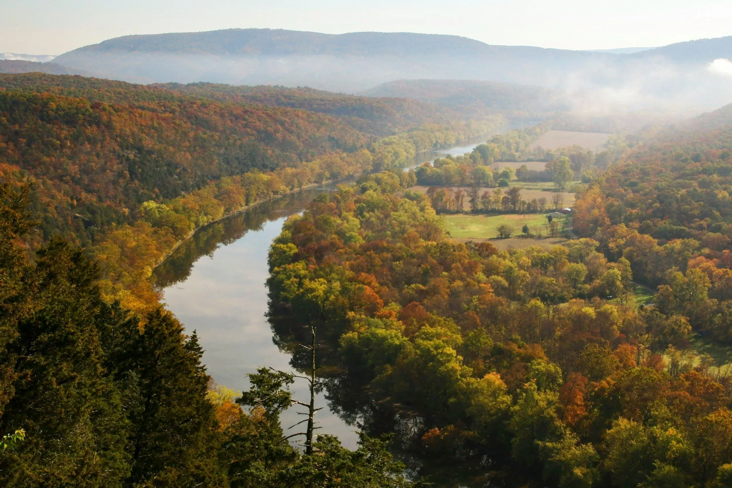 Aerial view of a winding river through an autumn-colored forest valley, farmland, and misty mountains