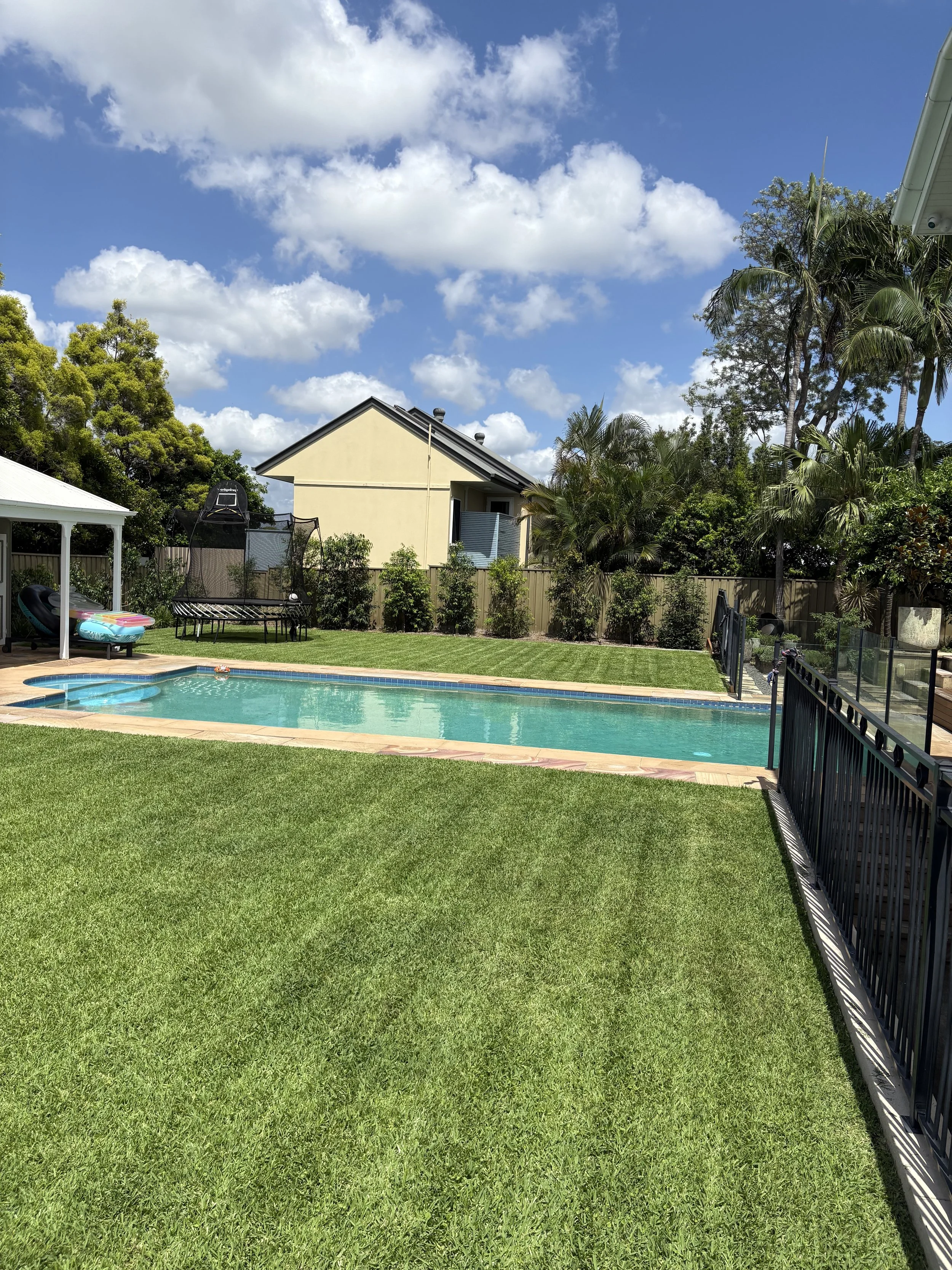 Backyard with a swimming pool, green grass, trees, and a fence under a partly cloudy sky.