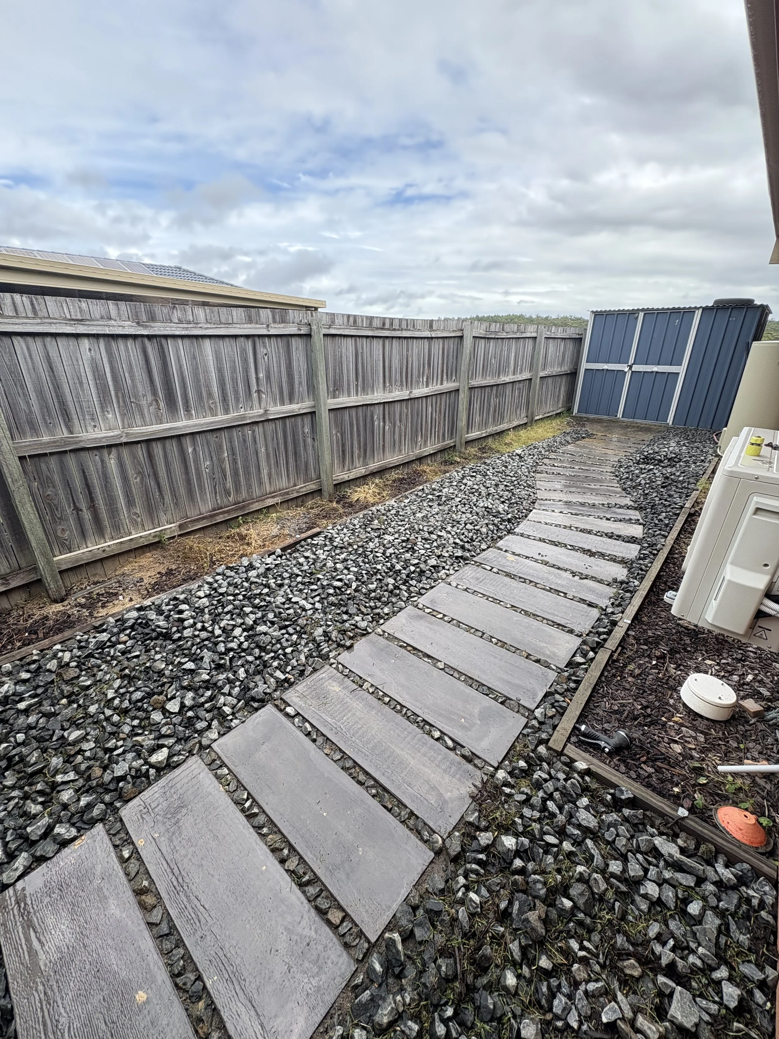 A backyard pathway with wooden stepping stones and gravel on either side, bordered by a wooden fence and a blue storage shed in the background.
