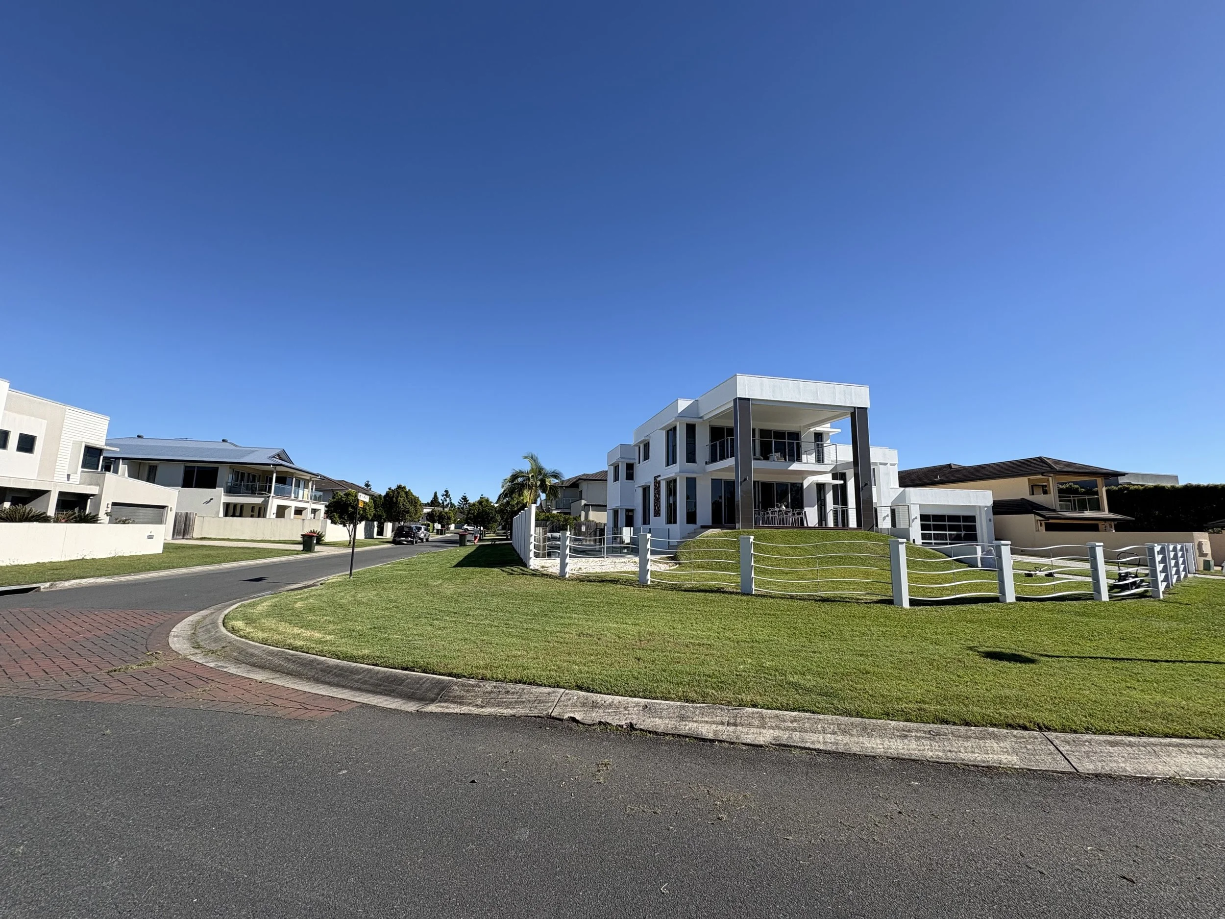 Modern two-story white house with large balconies, surrounded by a well-maintained lawn and a white fence, on a sunny day with a clear blue sky.