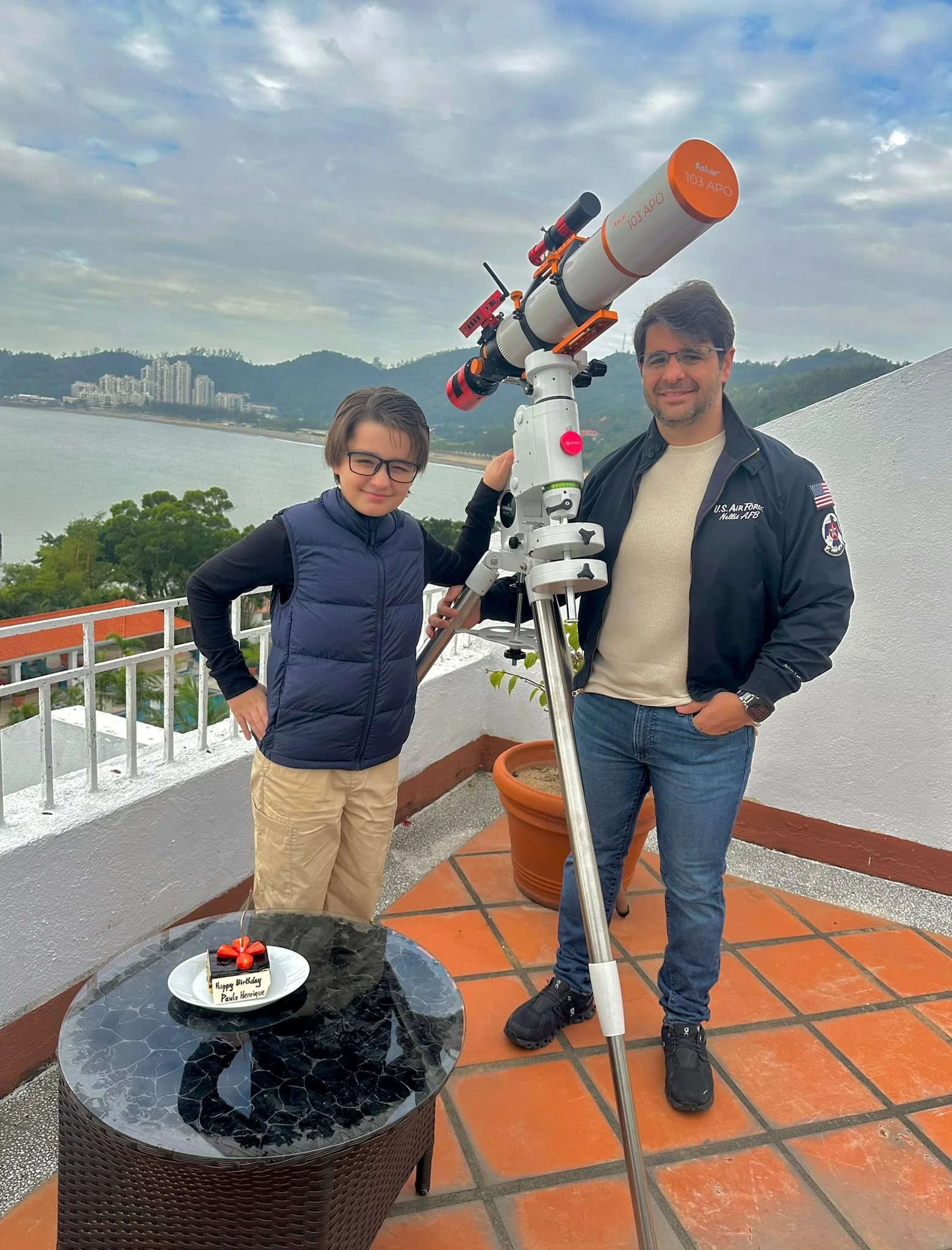 A boy and a man stand on a balcony, looking through a telescope. The boy is wearing glasses and a blue vest, the man is wearing a black jacket with U.S. Air Force patches. There is a small birthday cake on a black table in front of them, and a scenic view of a body of water, buildings, and hills in the background.