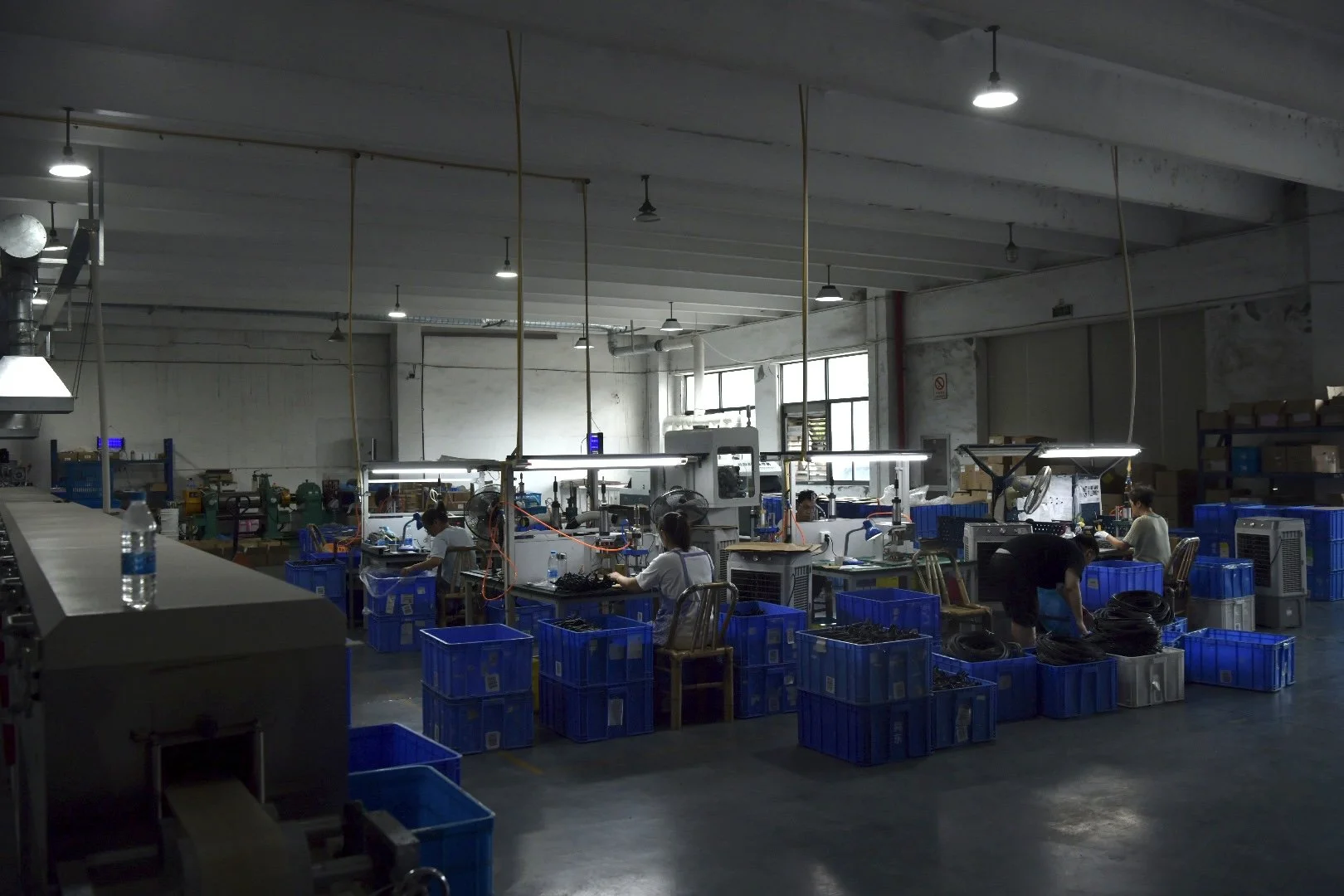 Workers in a factory assembling electronic components at workstations with blue storage bins, industrial lighting, and large windows in the background.