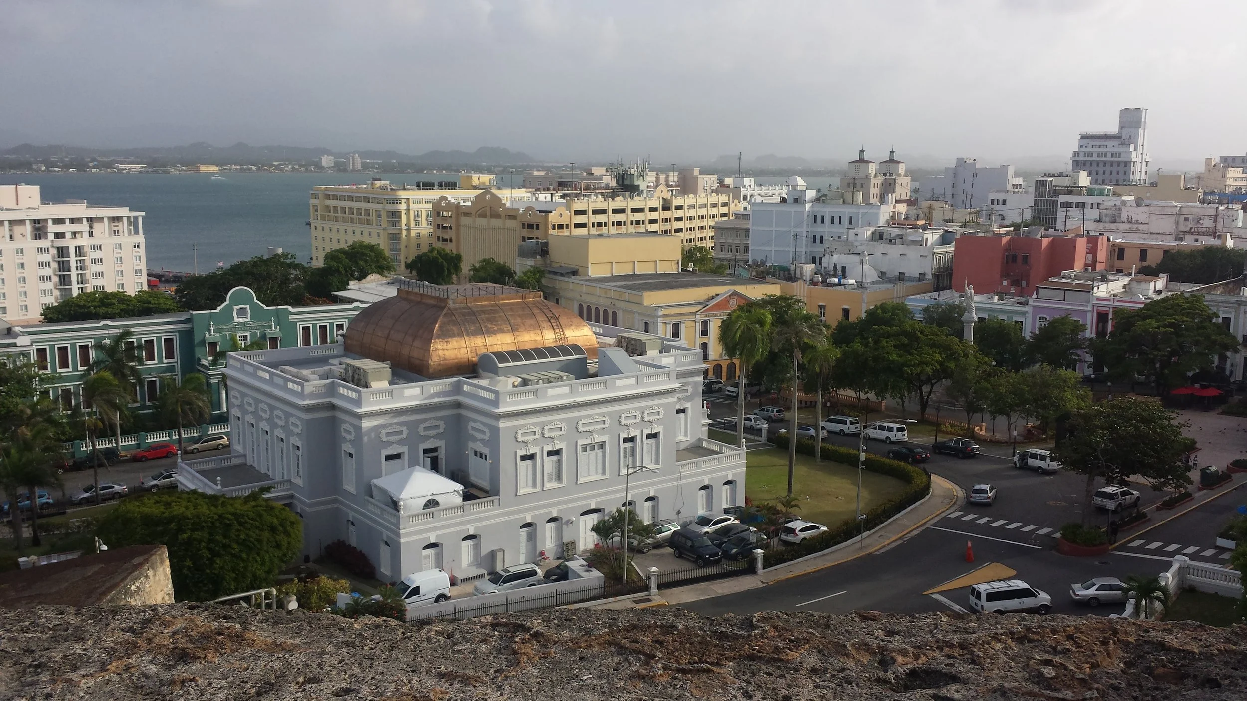 Cityscape with historic white building featuring a gold dome in the foreground, surrounded by trees, various multi-story buildings, a water body, and a distant horizon with mountains under a cloudy sky Bird's eye view of Puerto Rico