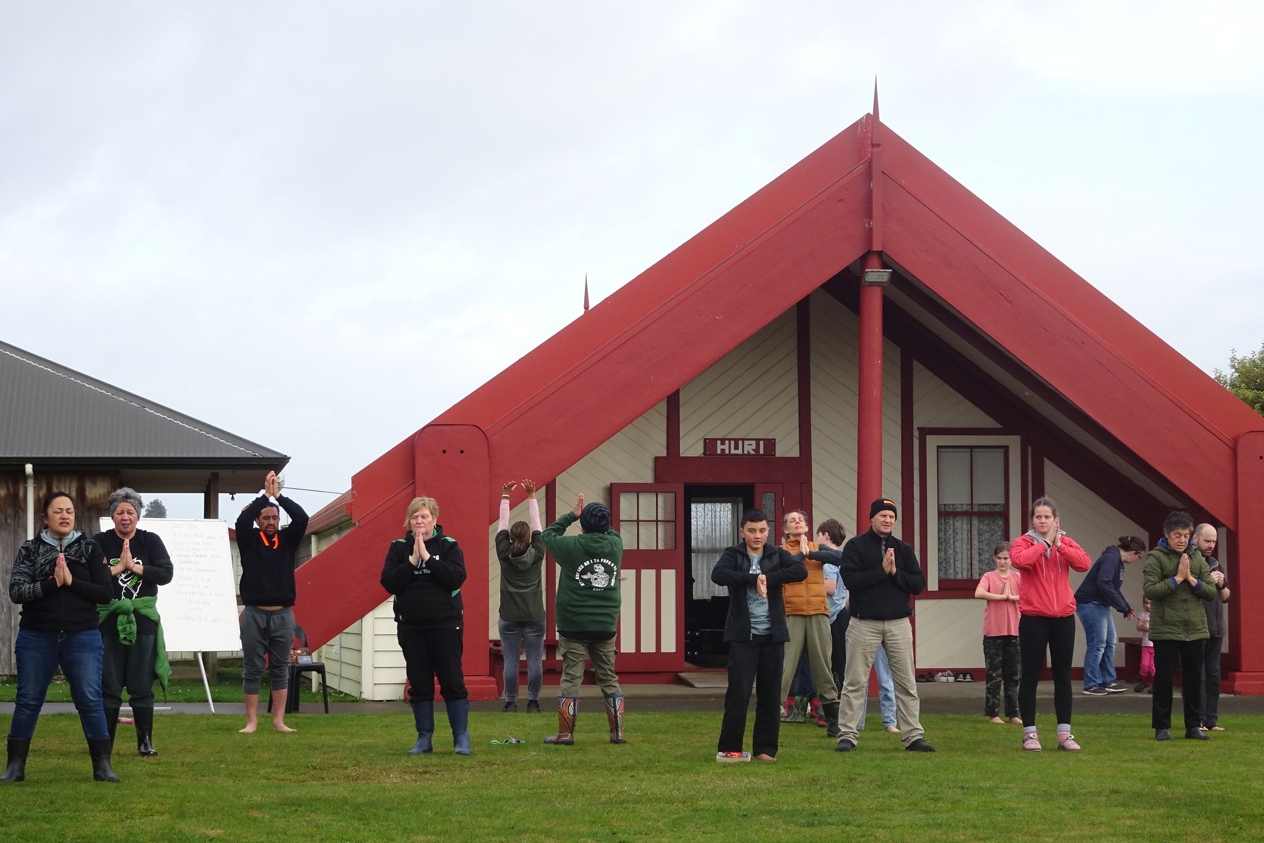 Group of people practicing yoga outdoors in front of a building with a red roof and beige walls, some with hands together in prayer pose, others doing stretching or standing poses.
