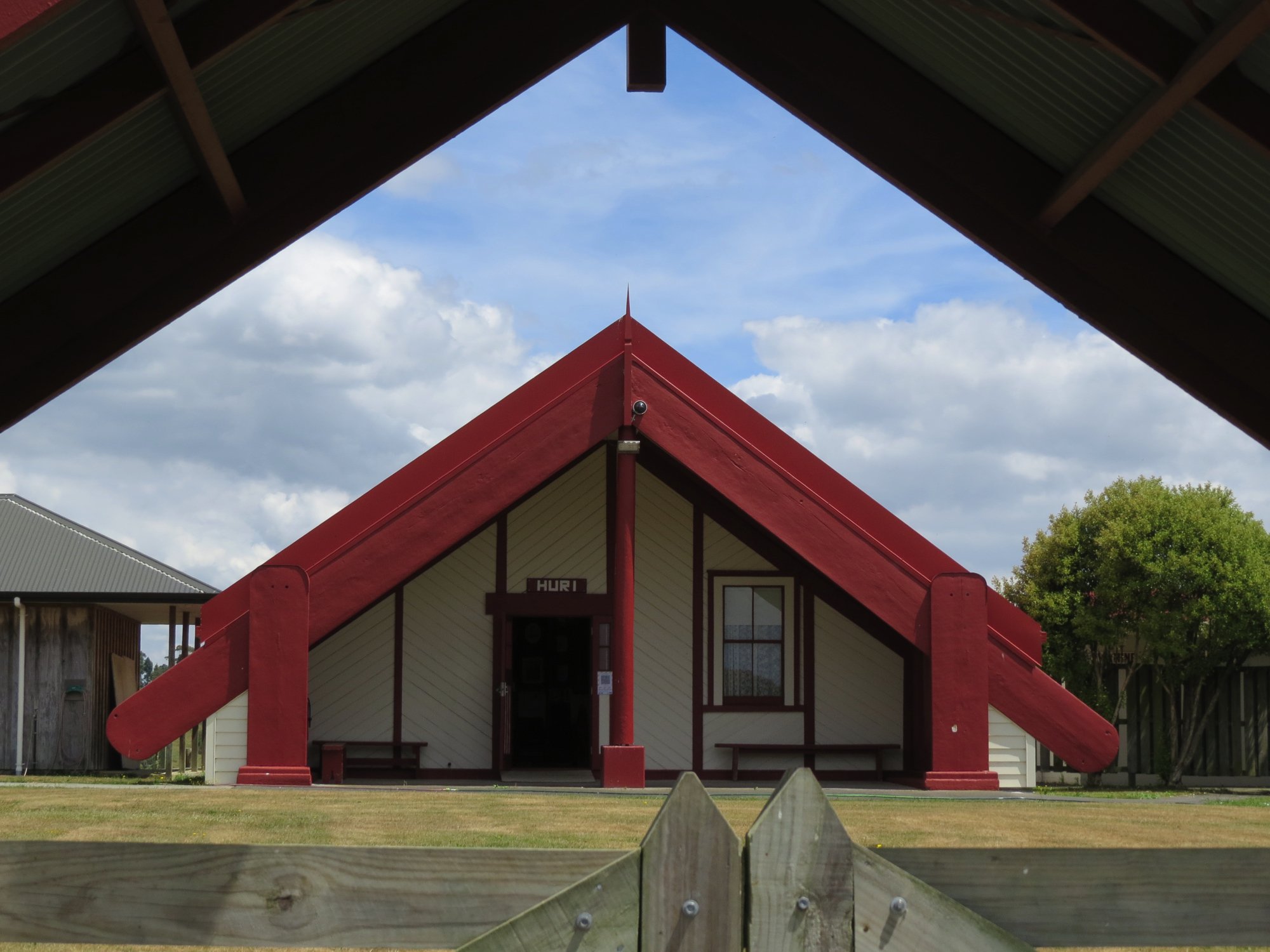 A building with a sharply peaked red roof, white walls with black trim, and a small porch with benches, viewed through a wooden gate and framed by two structures with green corrugated roofs.