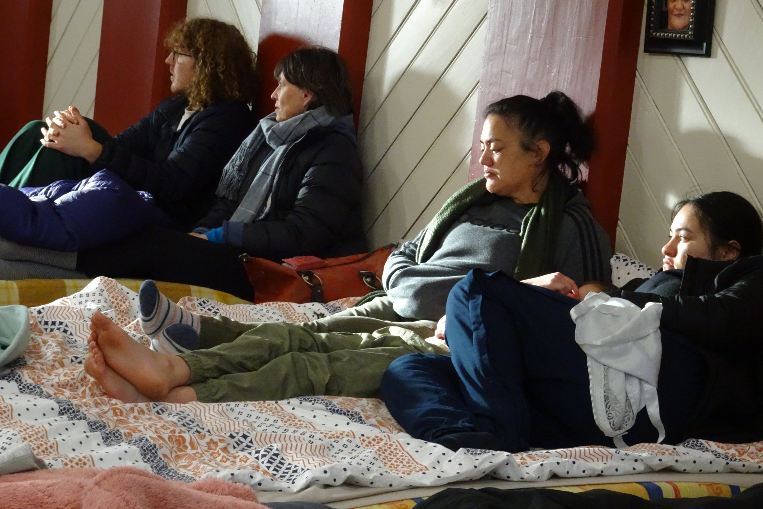 Several people are sitting on a bed in Huri, with some curled up with eyes closed, appearing to be resting or sleeping.