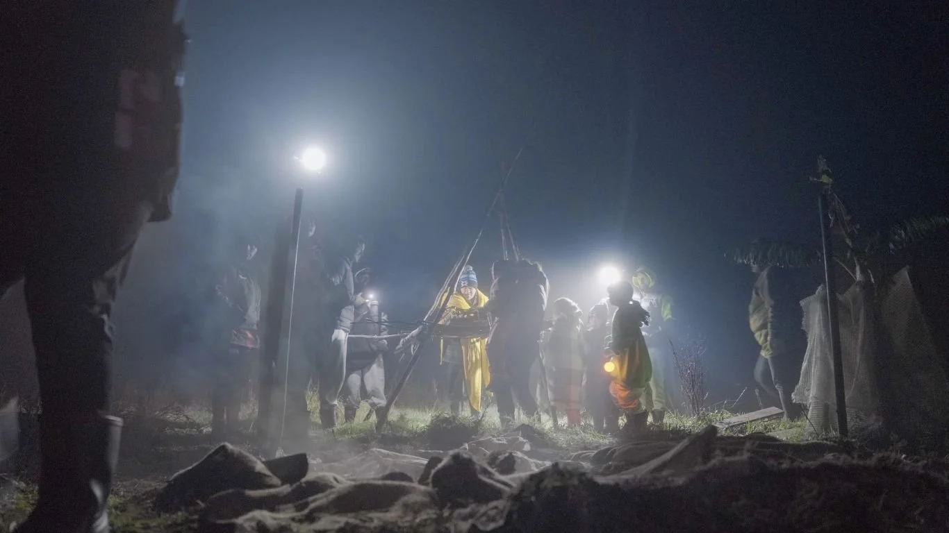 Several people gathered outdoors at night, illuminated by flashlights, with a bright moon in the sky and dry grass and rocks in the foreground.