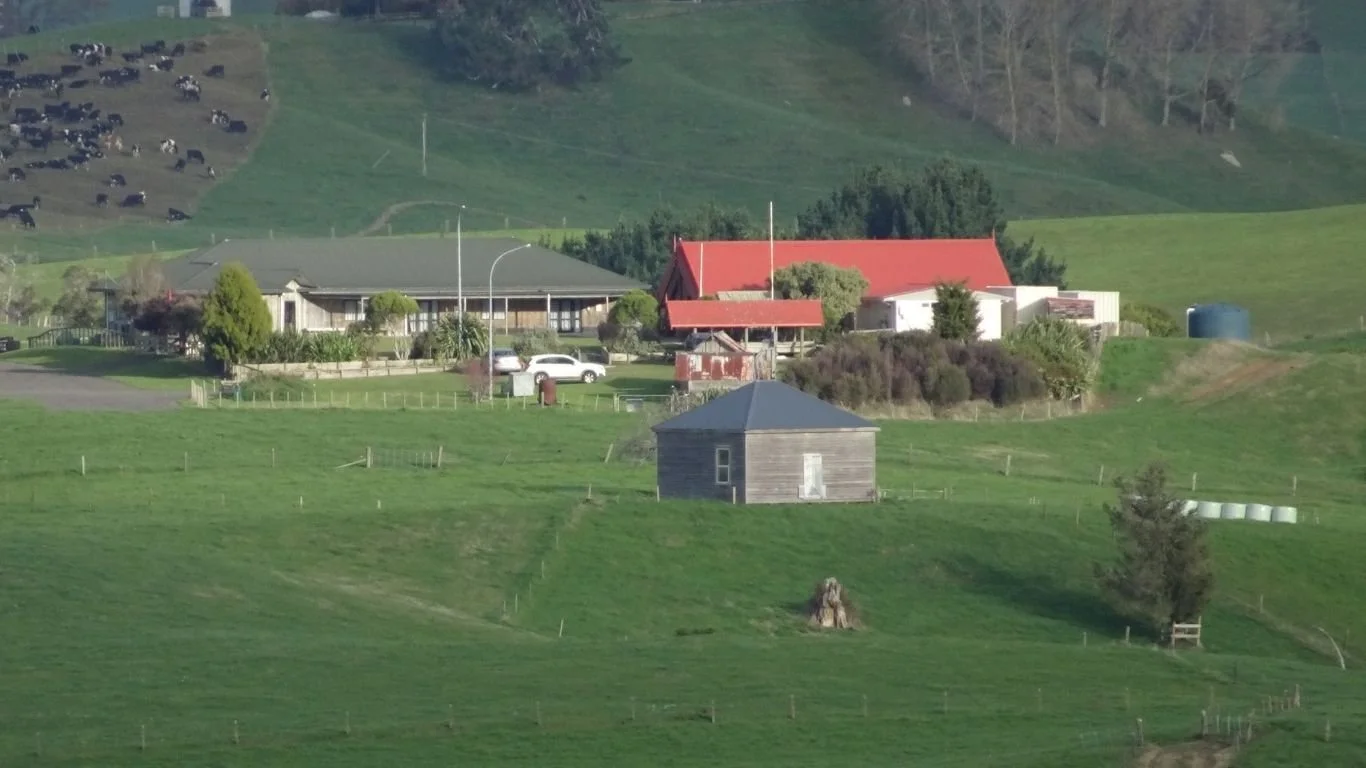 A rural landscape with green rolling hills, farm buildings, trees, cows grazing in the background, and a small wooden structure in the foreground.