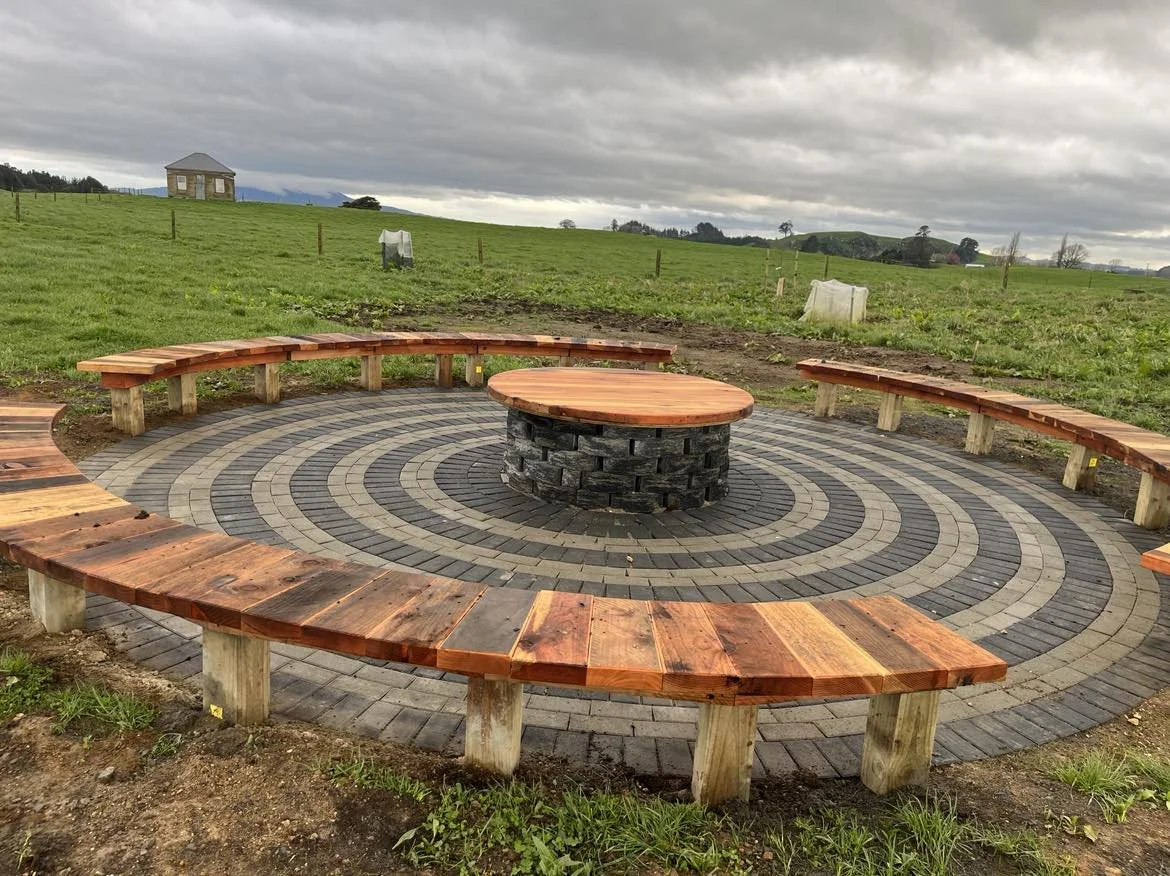 Circular outdoor seating area with wooden benches and a central table, surrounded by a brick pattern, in a green field under cloudy sky.