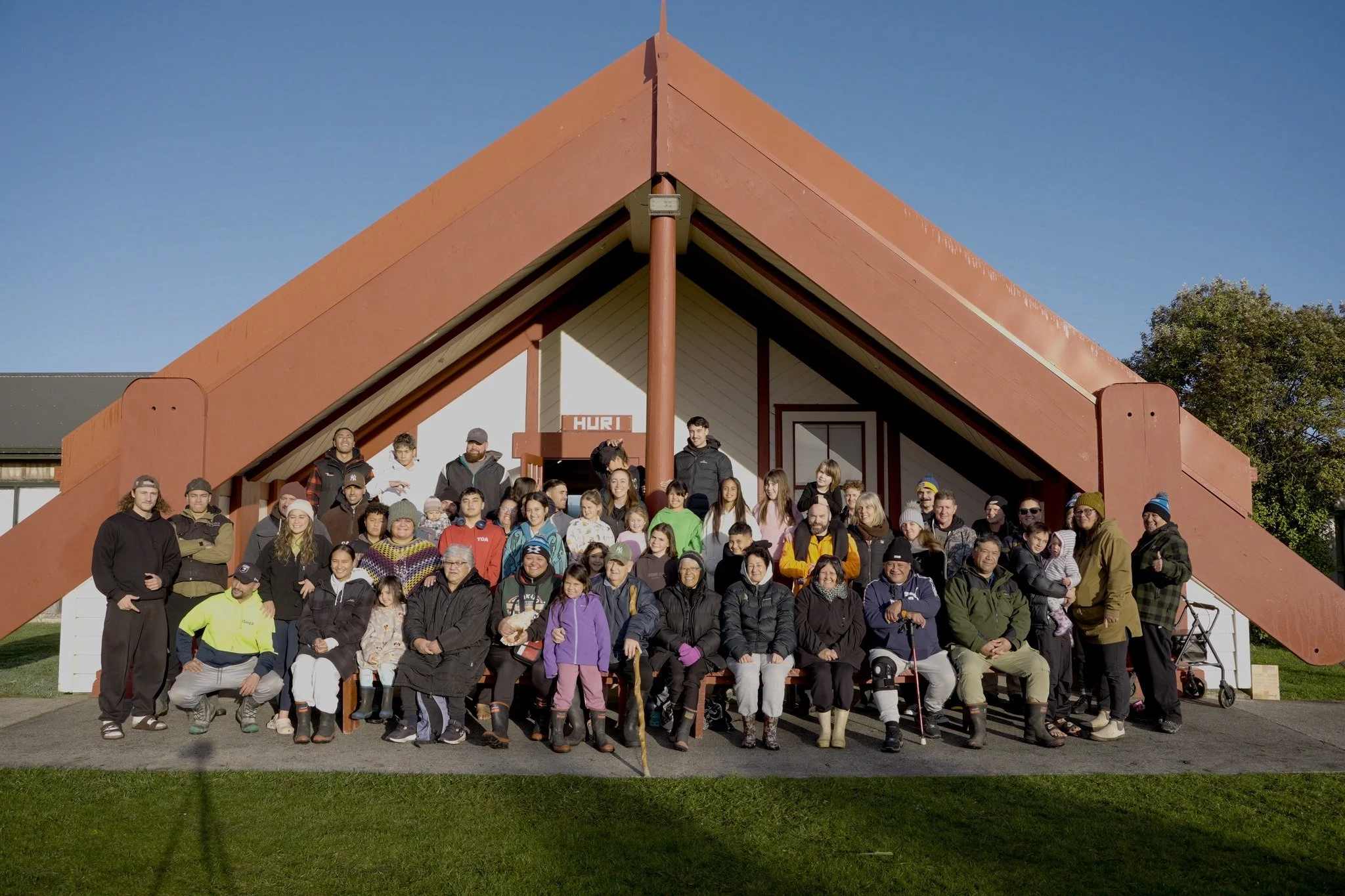A large group of Pikitū whānau gathered in front of a building with a steep red roof on a sunny day.