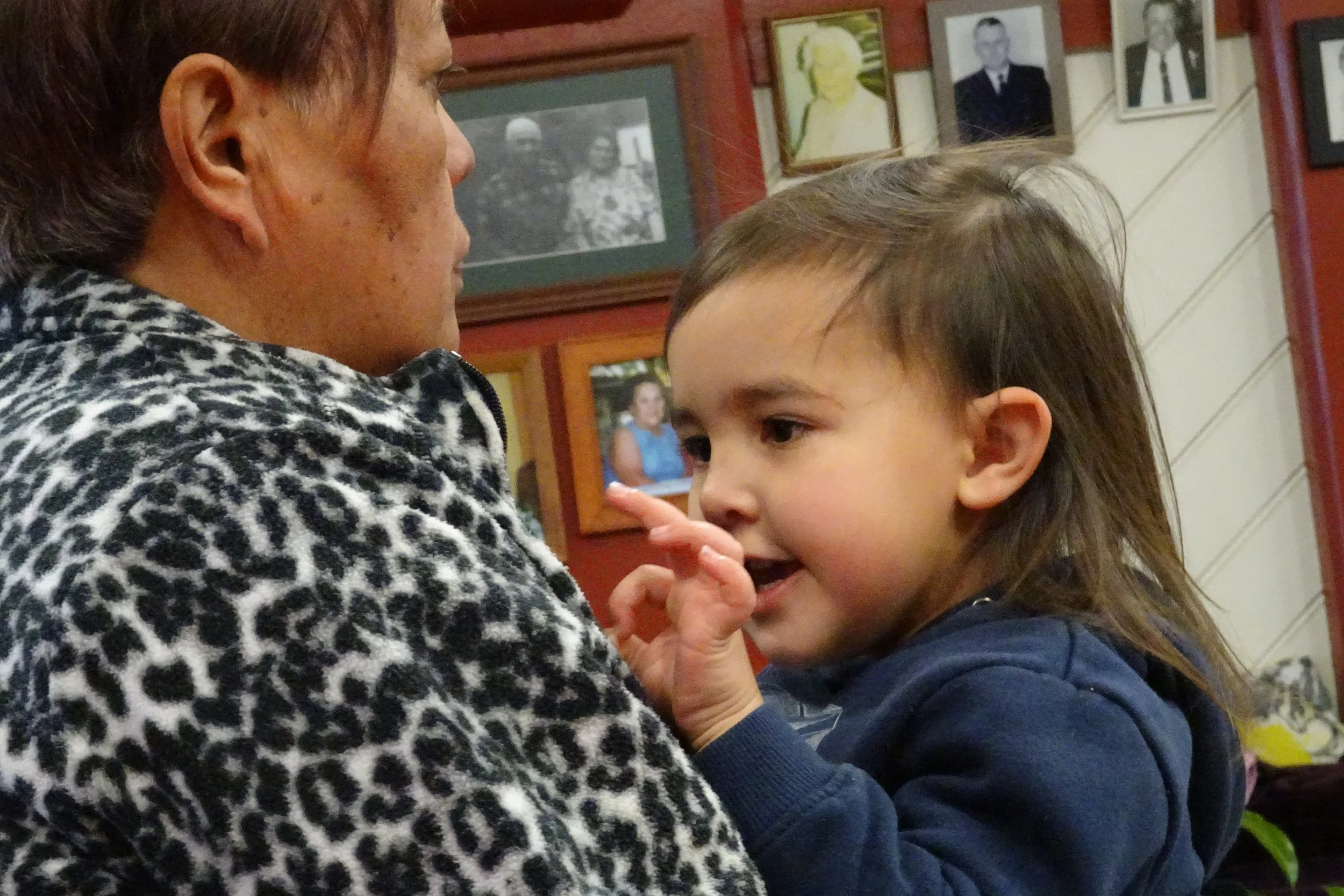 A young girl interacts closely with an older woman indoors, with framed photos on a red and cream wall in the background.