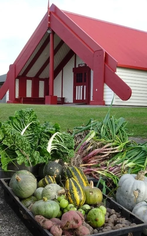 A basket of freshly harvested vegetables, including lettuce, squash, green tomatoes, and sweet potatoes, in front of a red barn with a covered porch.