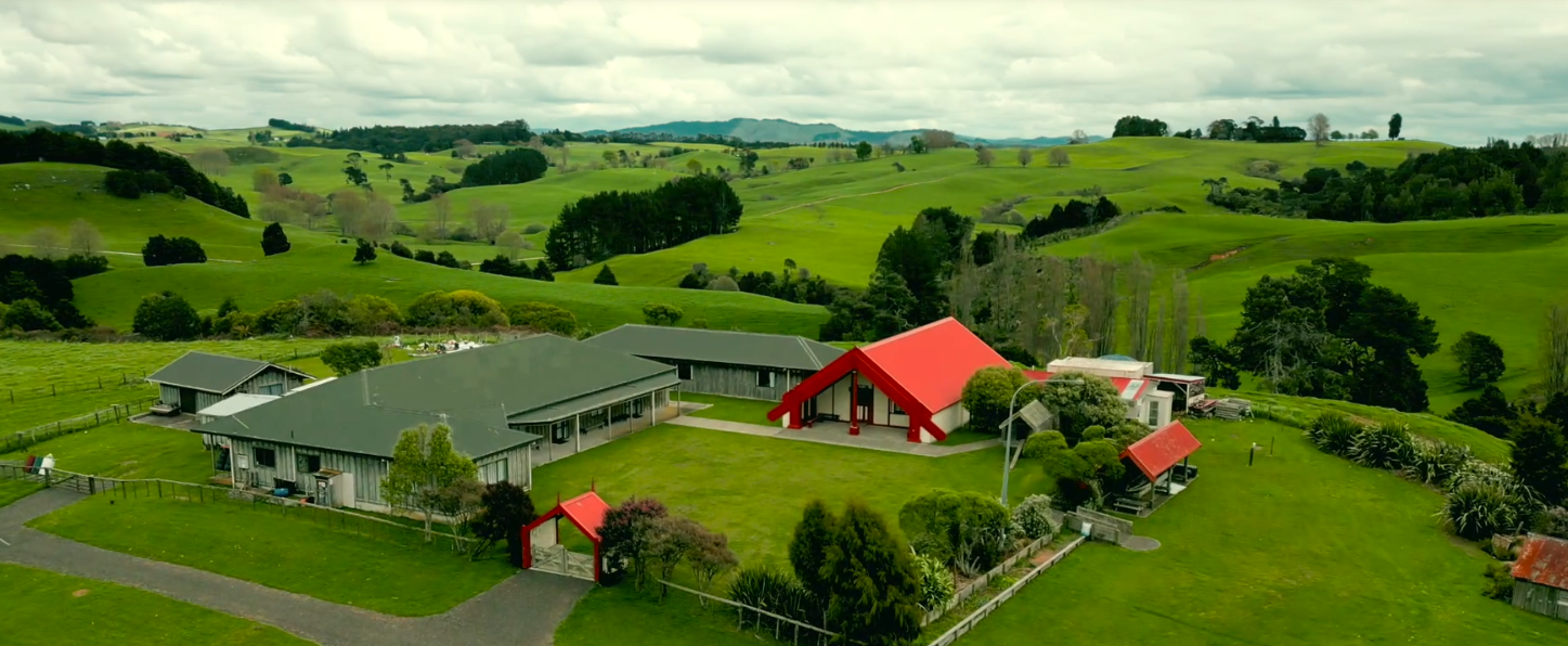 A scenic aerial view of a marae with several buildings, lush green rolling hills, trees, and cloudy sky in the background.