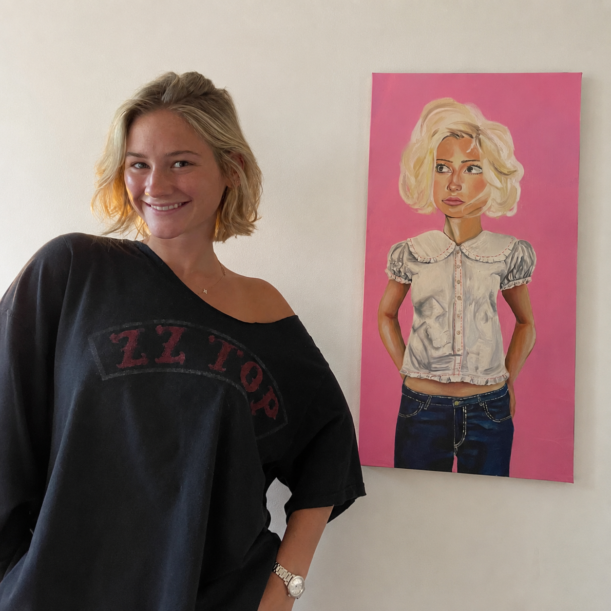A young woman with blonde hair smiling and wearing a black shirt standing next to a colorful portrait of another woman with blonde hair, wearing a light-colored blouse and jeans, with a pink background.