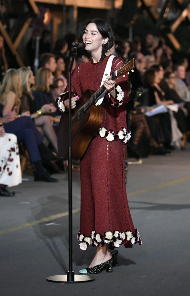 A woman in a maroon dress with floral embroidery, playing an acoustic guitar and singing into a microphone at an indoor event with an audience in the background.