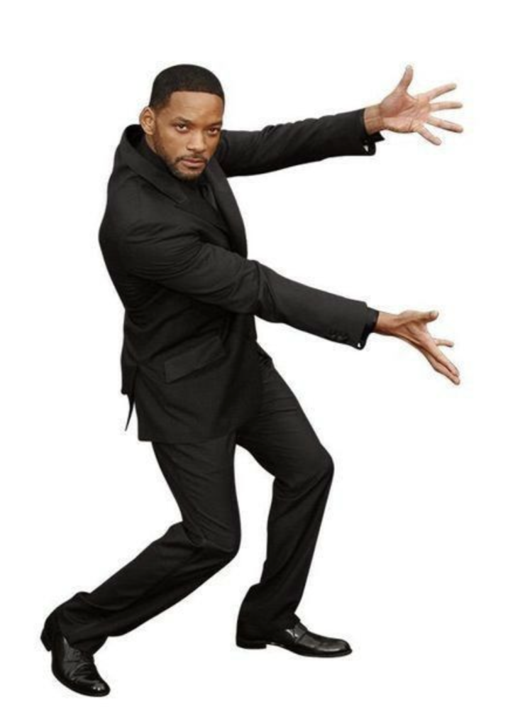 A man in a black suit posing with one arm extended forward and the other arm extended sideways, standing with legs apart against a plain white background.
