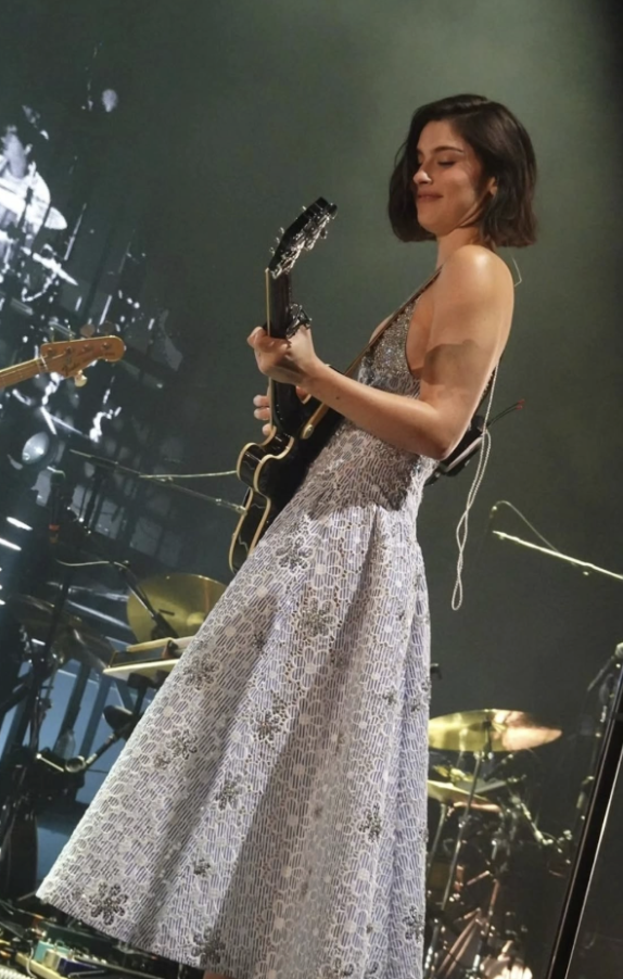 A woman playing an electric guitar on stage, wearing a patterned sleeveless dress, with a drum set and other musical equipment in the background.