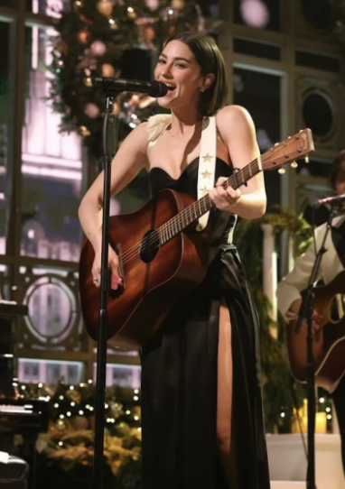 A woman singing into a microphone and playing an acoustic guitar during a festive indoor performance, with Christmas decorations in the background.
