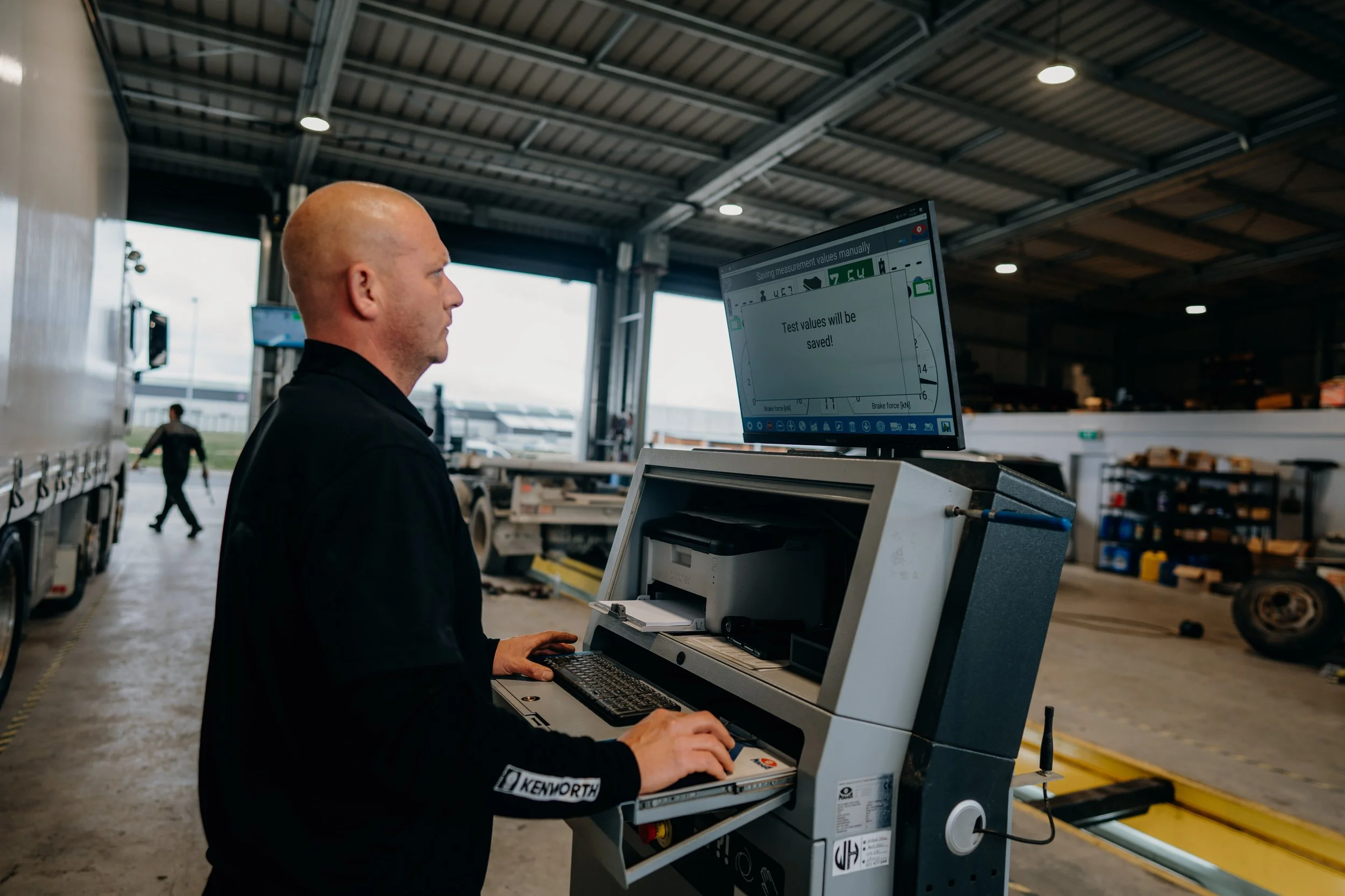 A man with a bald head and wearing a black shirt with the Kenworth logo is working on a computer terminal inside a large industrial warehouse. In the background, there are other people, storage shelves, and equipment.