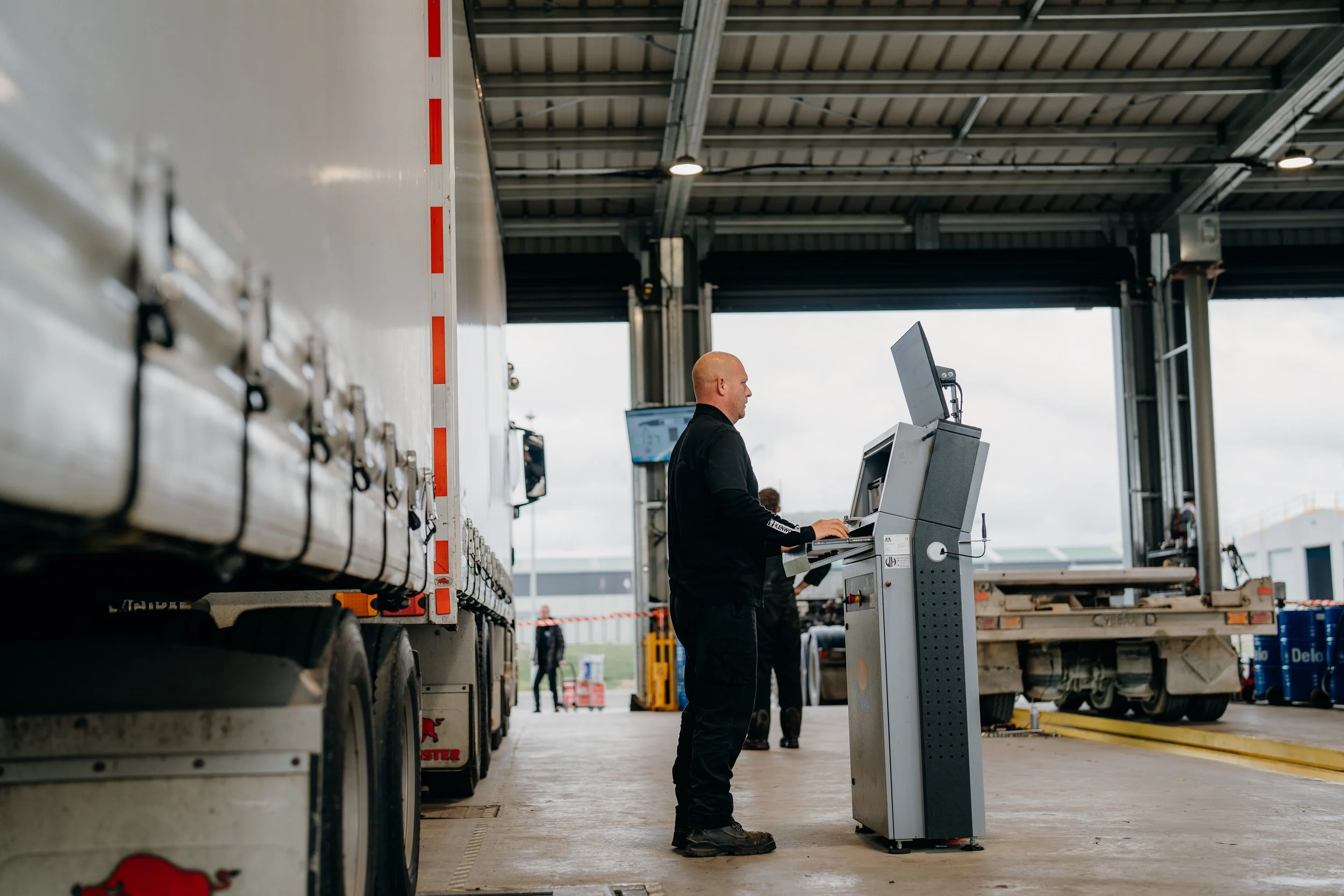A worker standing at a checkout terminal in a warehouse, with trucks and stored goods visible in the background.