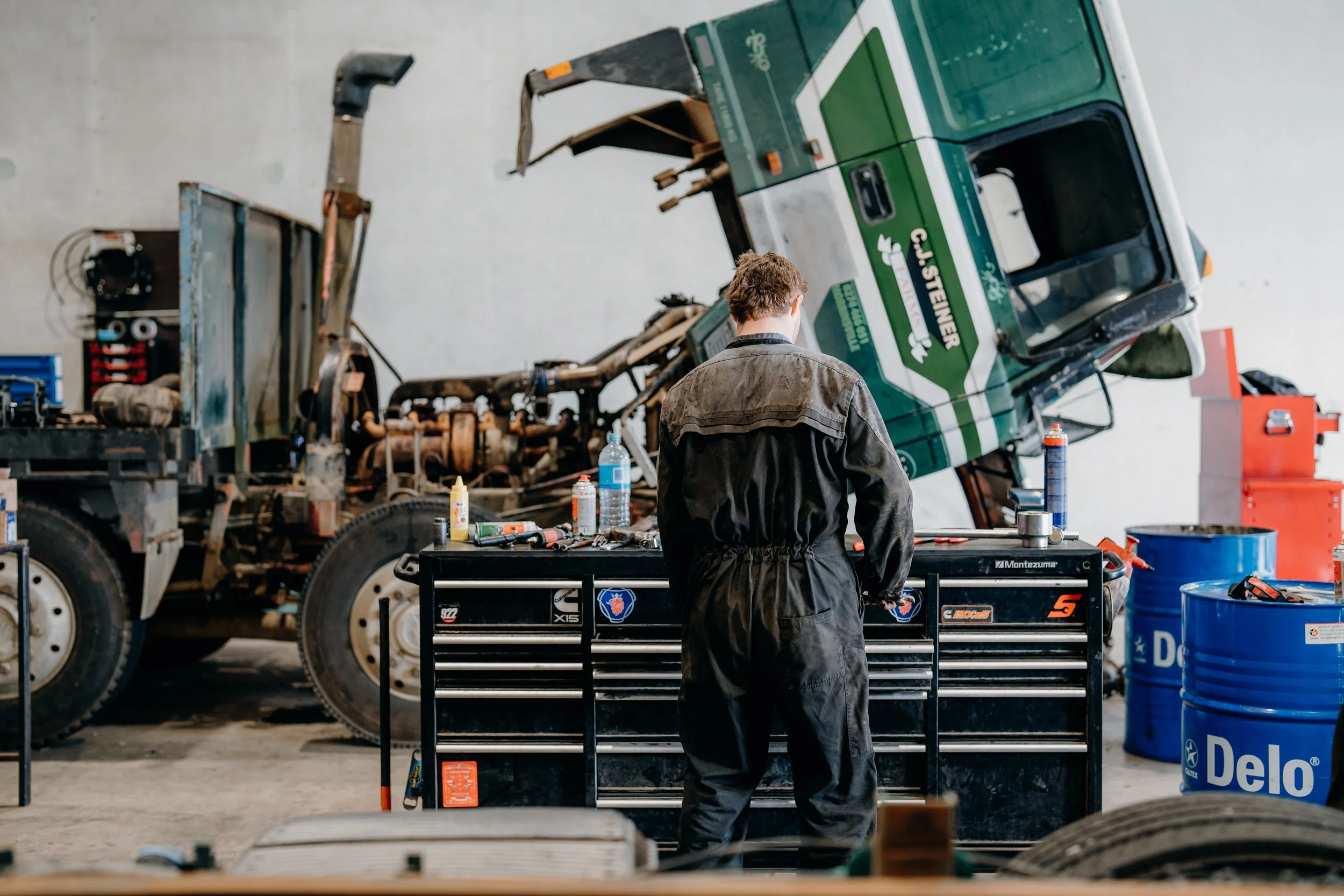 A person with short hair and wearing a black mechanic jumpsuit stands at a black workbench with tools, in an auto repair shop. Behind them is a large green tow truck with an extended hydraulic lift arm, and various tools and supplies are scattered around.