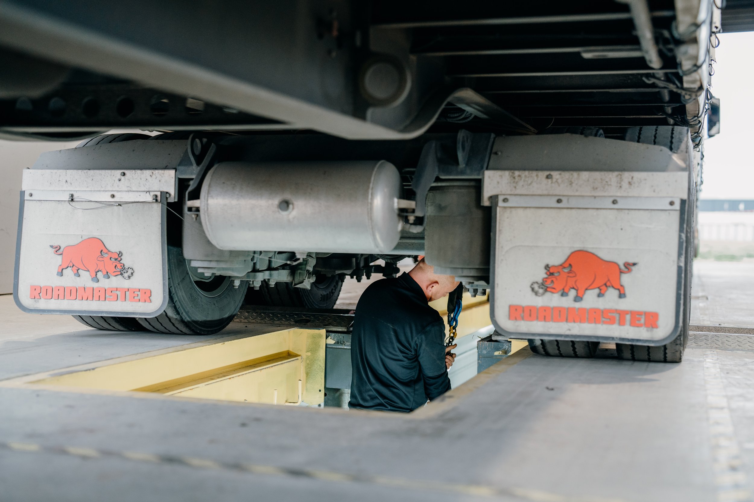 A person working underneath a large Roadmaster truck, with the focus on the truck's underside and wheels, and the worker's head partially visible.