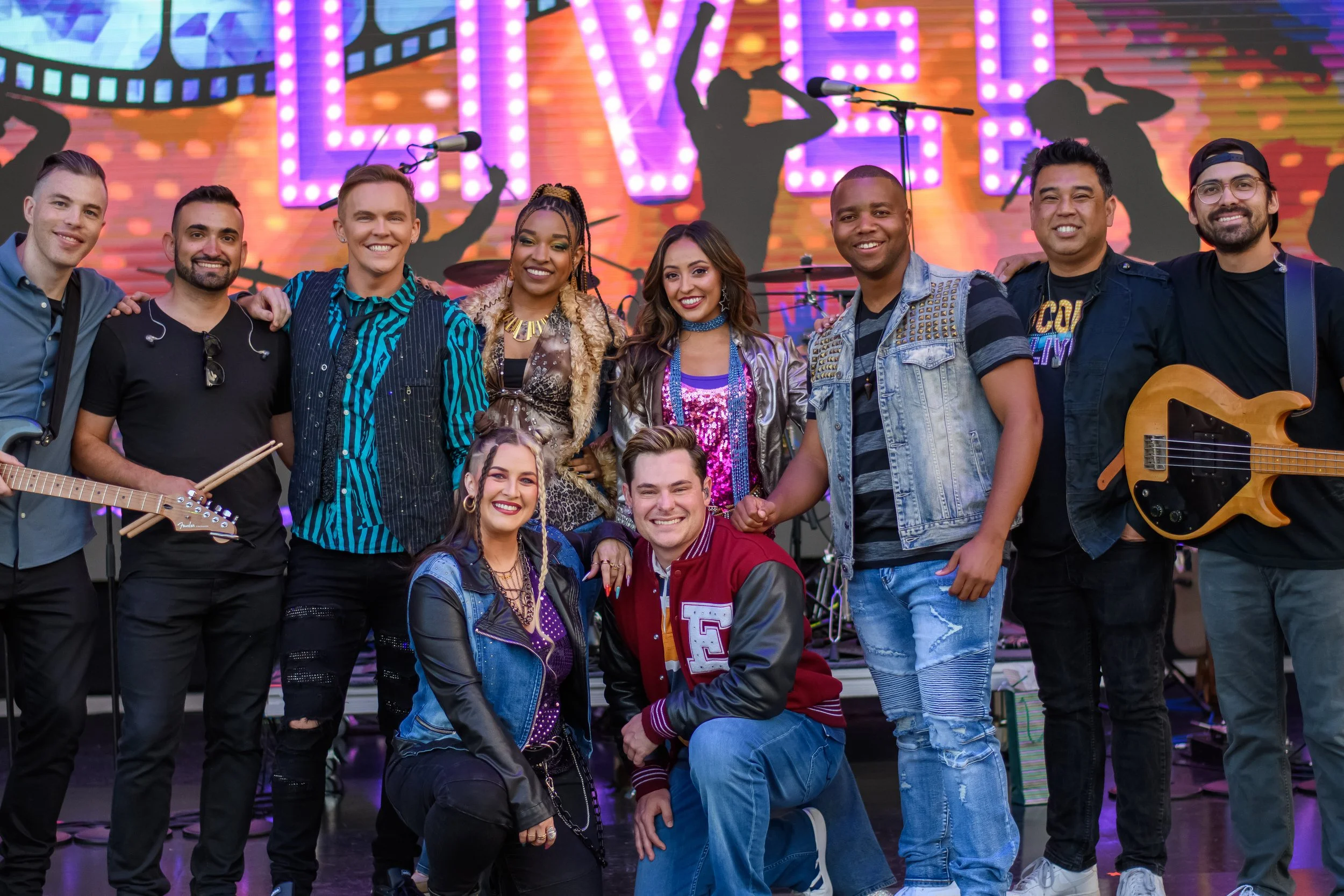 Group of nine people posing on a stage with musical instruments and colorful background. Some are holding guitars, and a drum set is visible behind them.