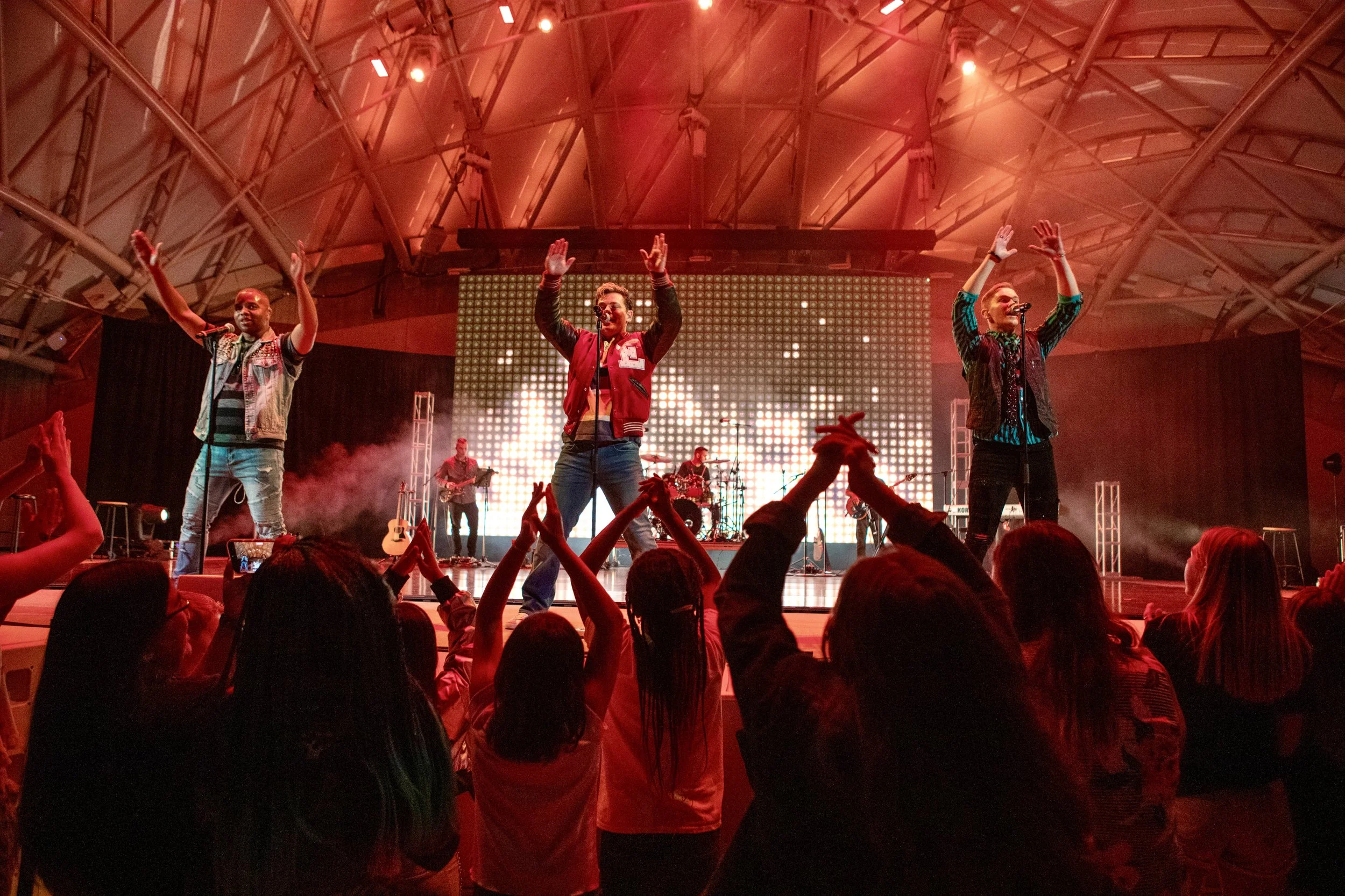 Musicians performing on stage at concert, audience raising hands, colorful lighting, large LED screen in background