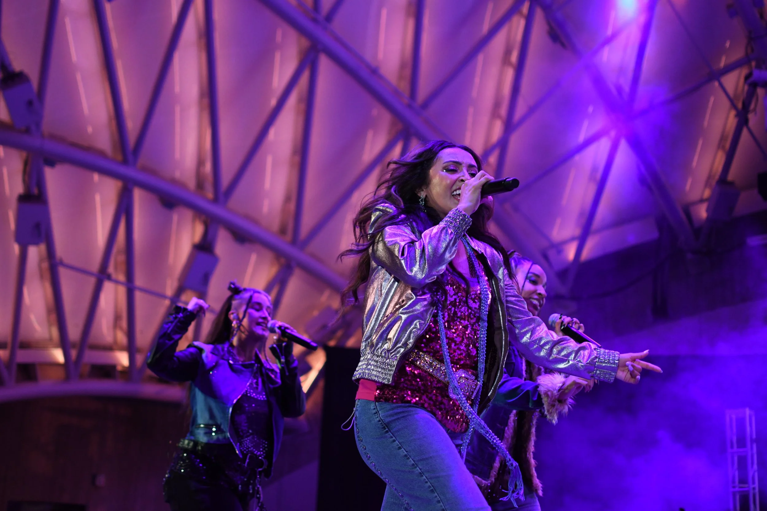 Four female singers perform on stage under purple lighting, with one singing into a microphone and others dancing.