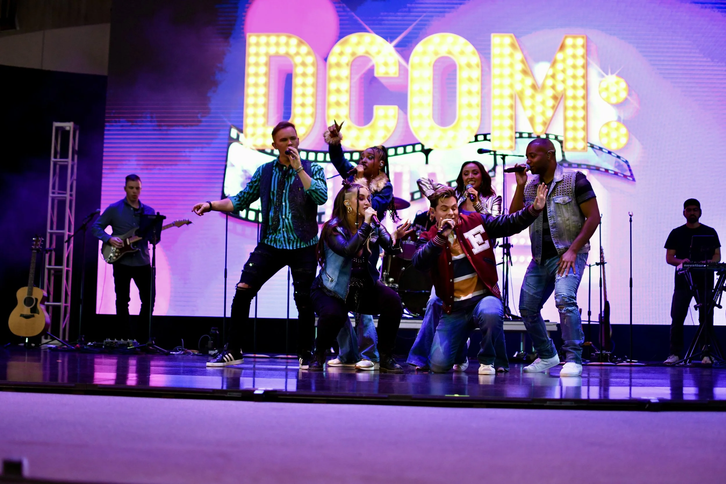 A group of seven young performers singing and dancing on stage with a colorful DC Comics-themed background and live band members playing instruments.