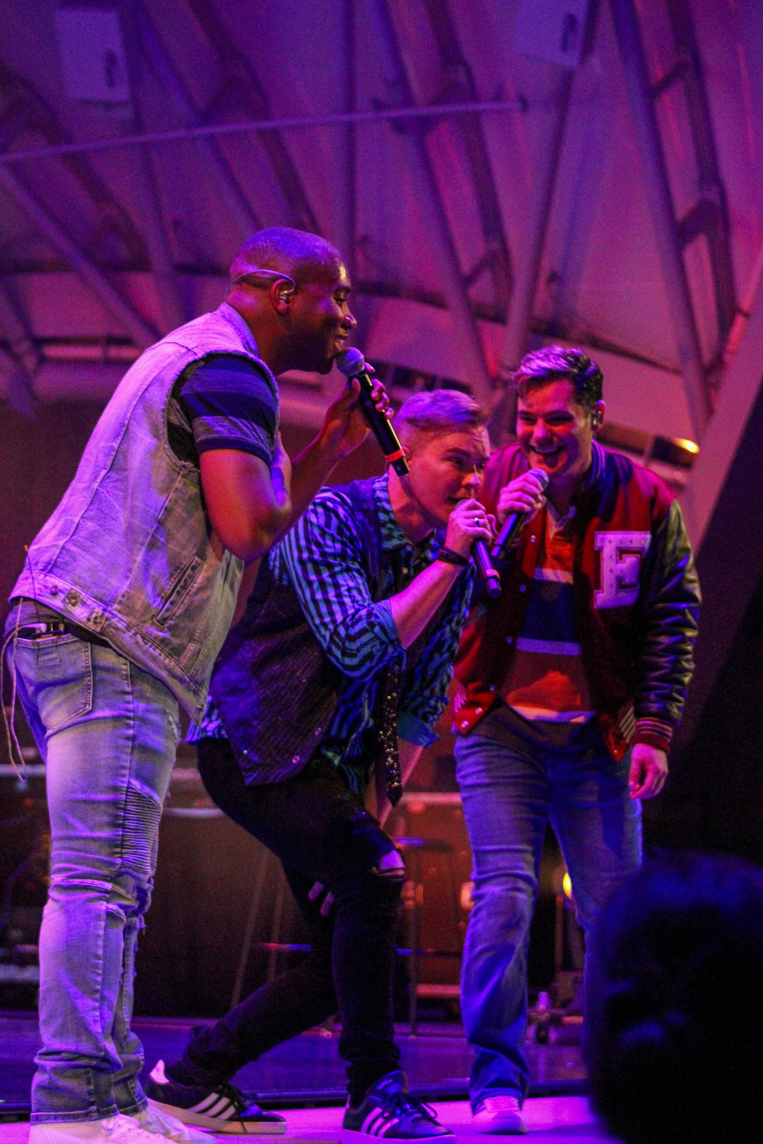 Three young men singing into microphones on stage with colorful lighting and a metal structure background.