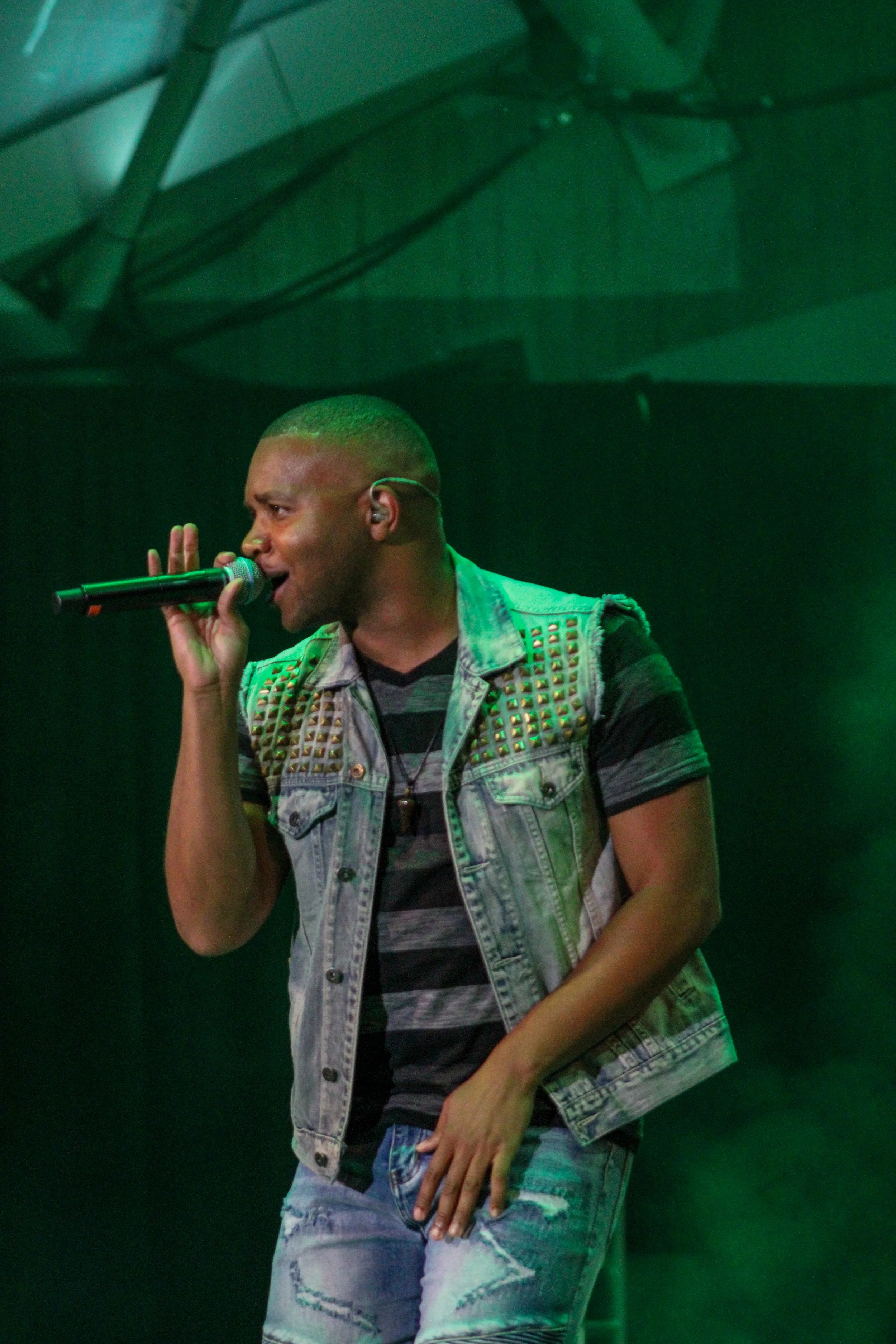 Male singer with short hair wearing a studded denim vest and ripped jeans, singing into a microphone on stage with green lighting.