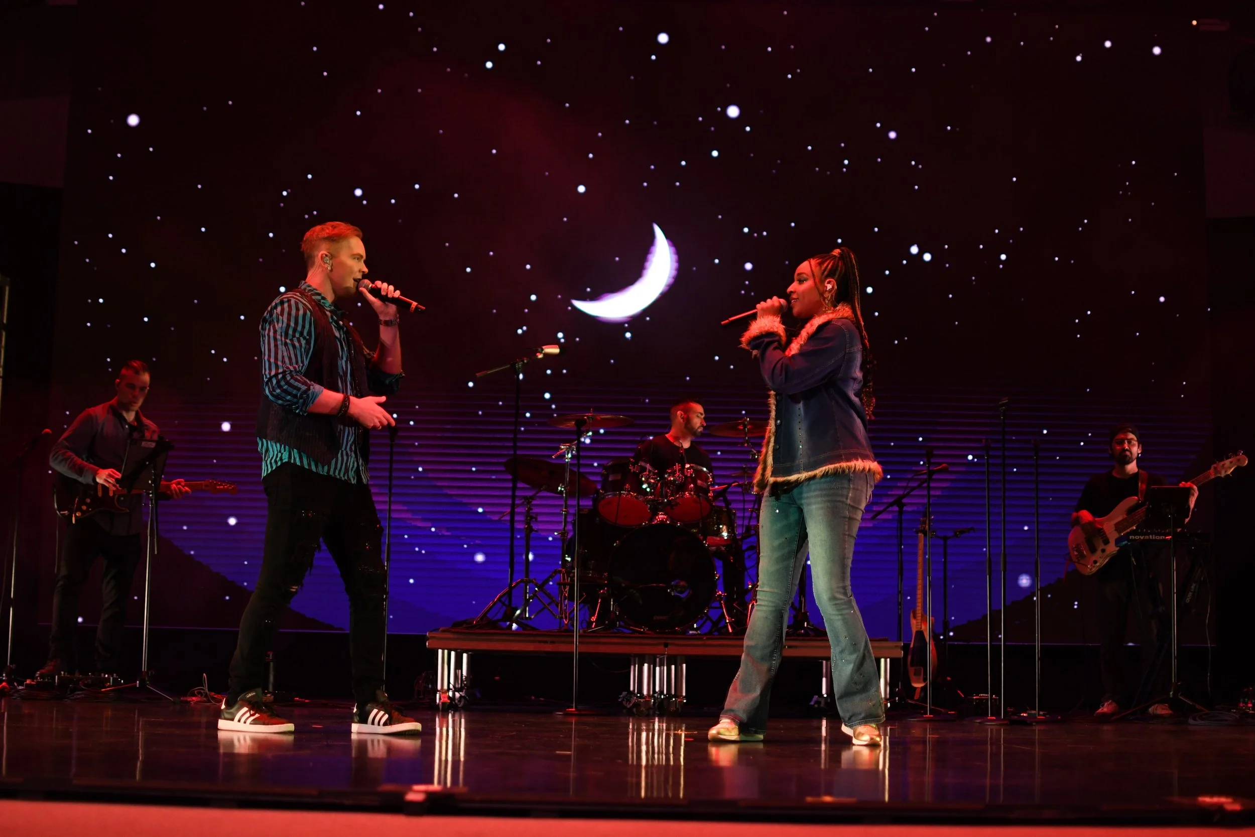 A band performs on stage with two singers holding microphones, a drummer, and two guitarists, against a backdrop of a starry night sky with a crescent moon.