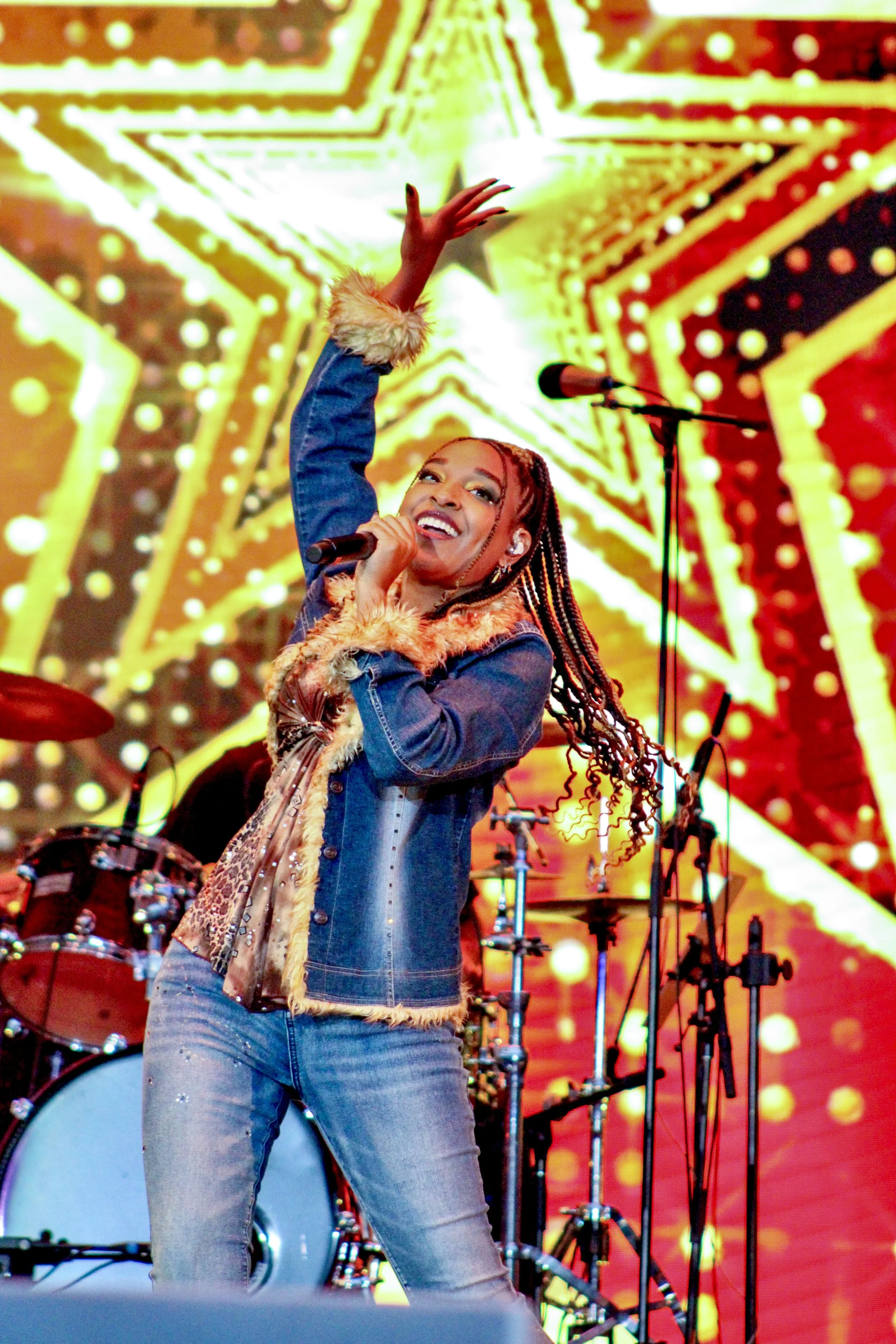 Performer singing into a microphone on stage, with a large, illuminated star graphic in the background and musical instruments visible behind her.