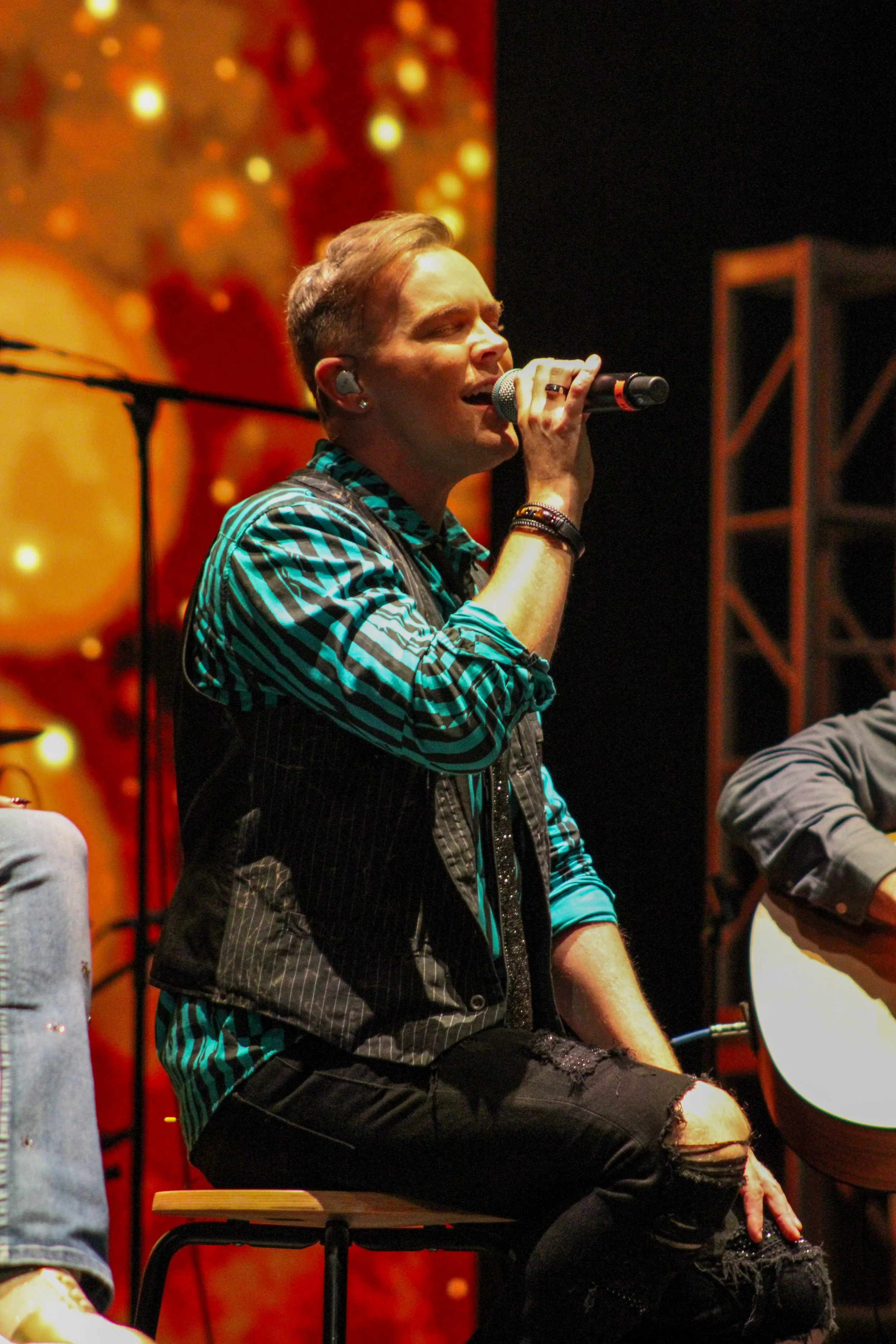 A person with short hair singing into a microphone while sitting on a stool, with a Christmas-themed background of golden lights and ornaments.