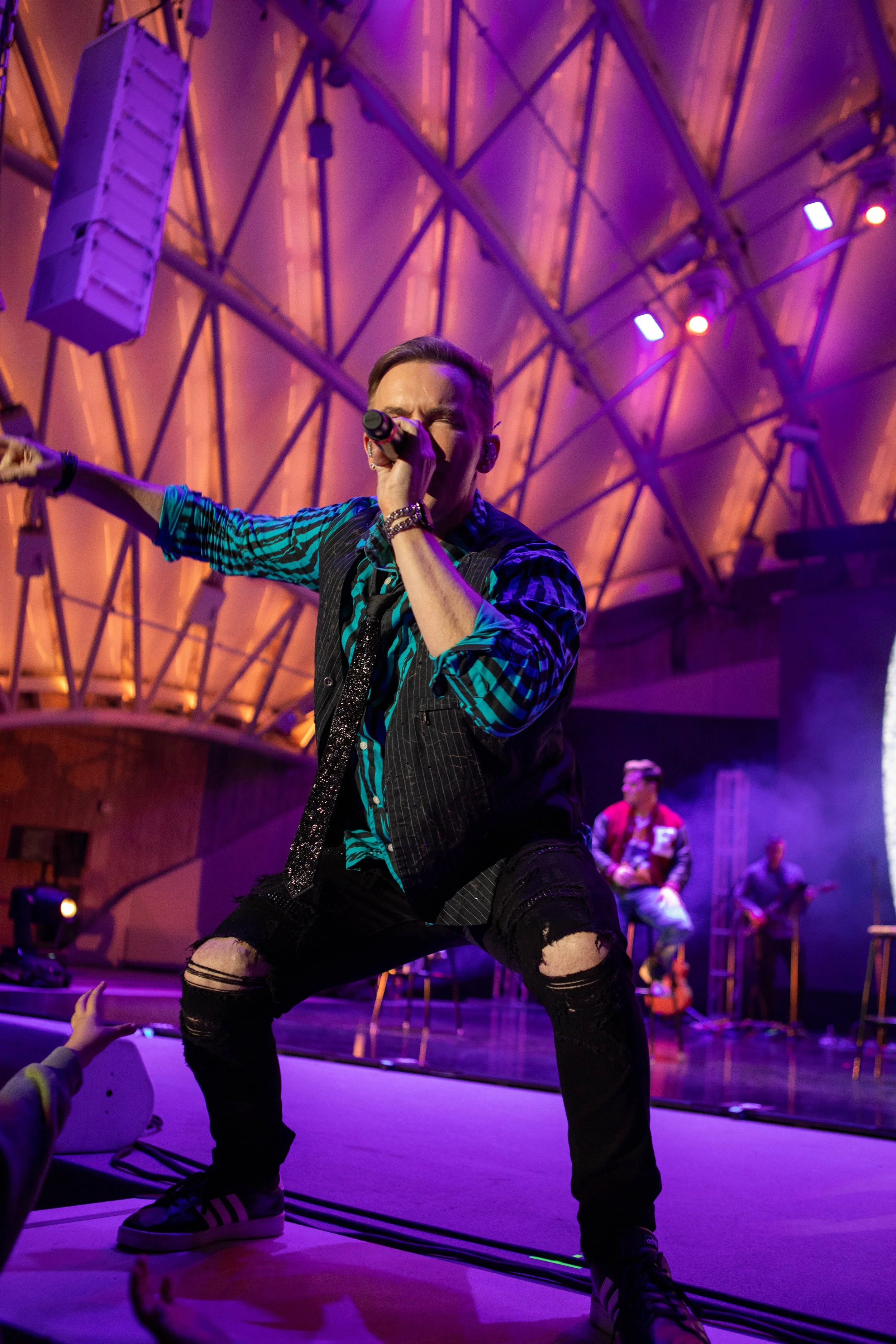 A male singer performing on stage with a microphone, wearing a striped shirt, a vest, and ripped jeans, with a band in the background. The stage has purple and orange lighting.