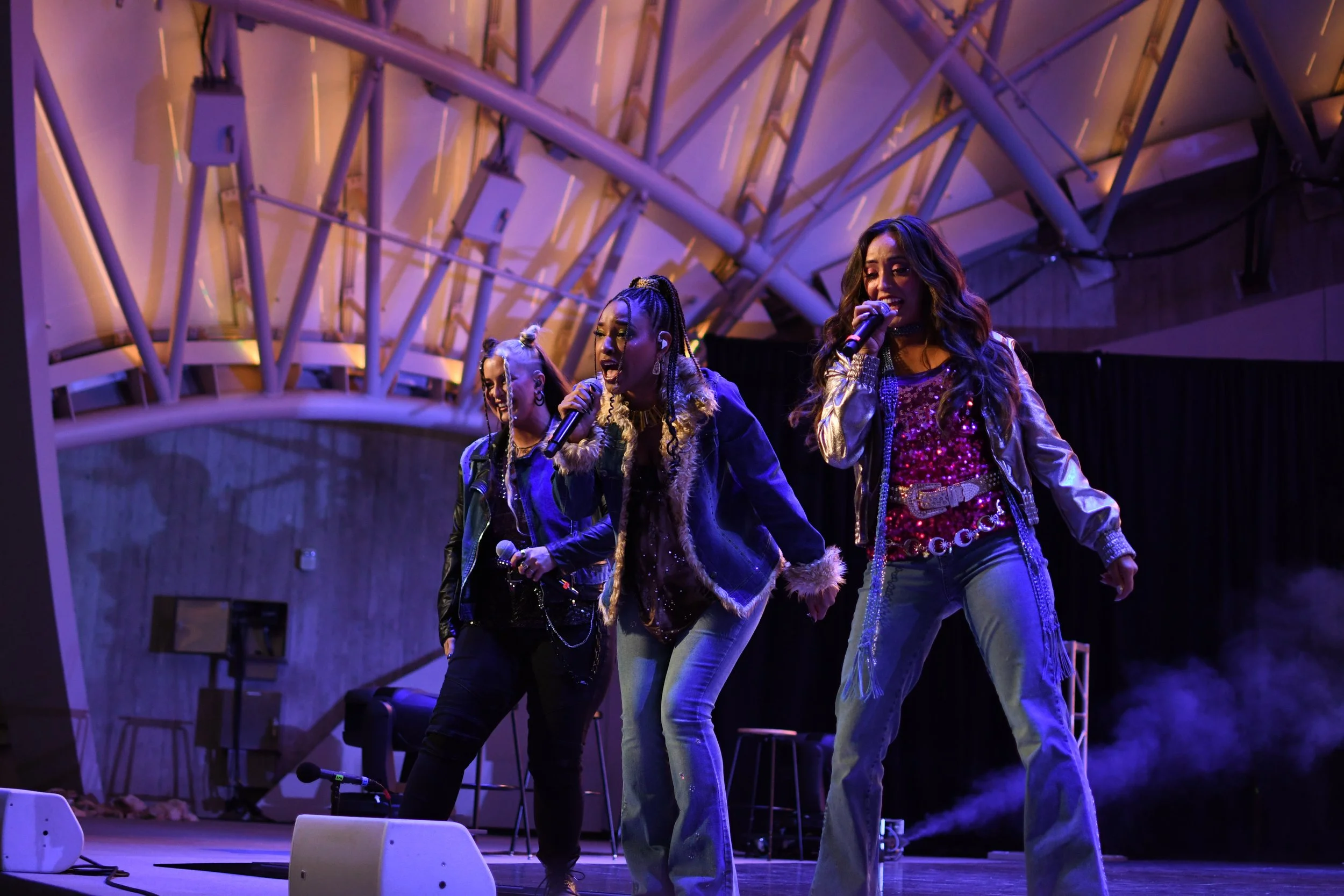 Three female singers performing on stage at a concert, holding microphones, with a modern architectural ceiling in the background.