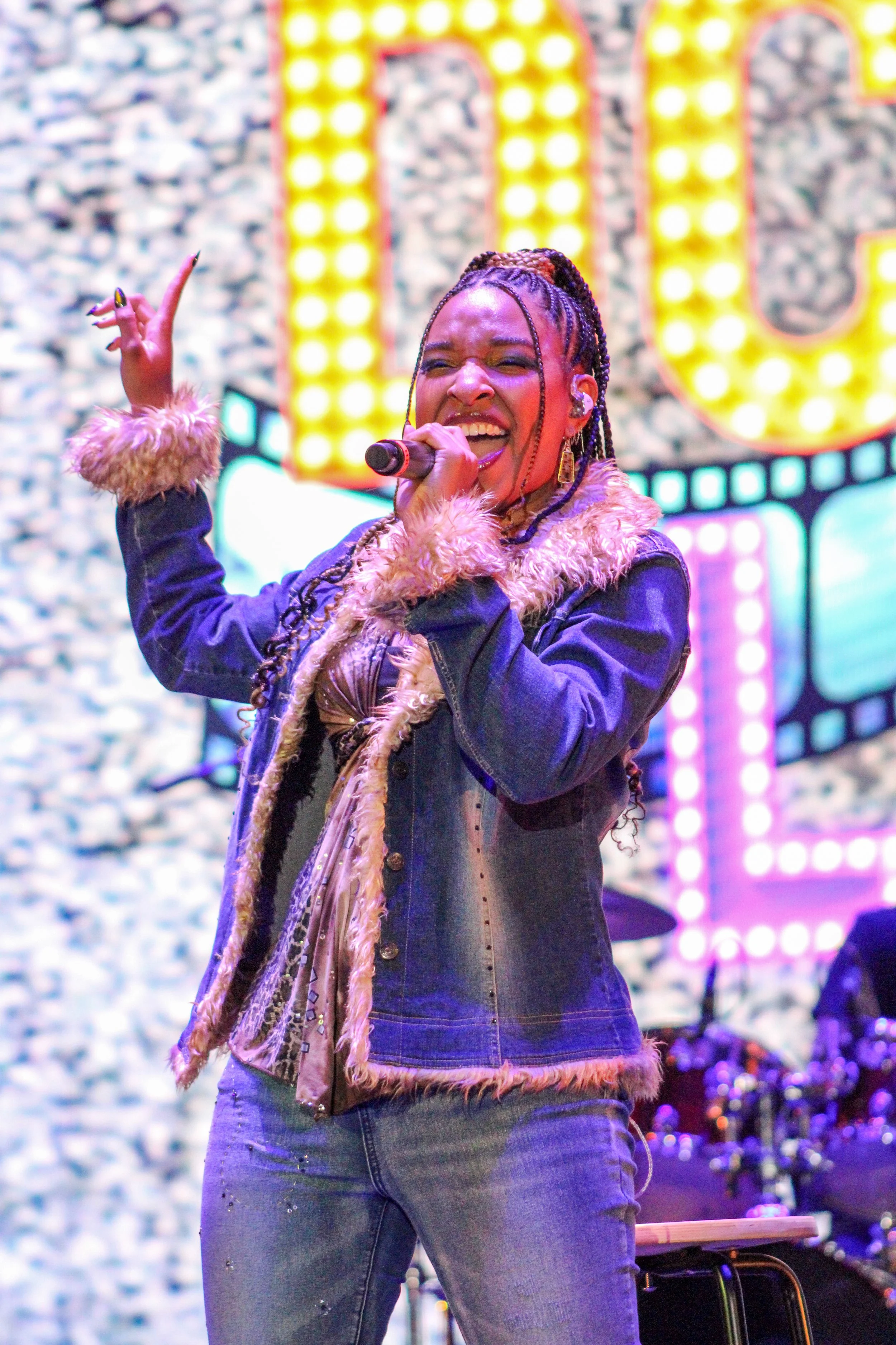 A woman singing into a microphone on stage with colorful lighting and a patterned background.