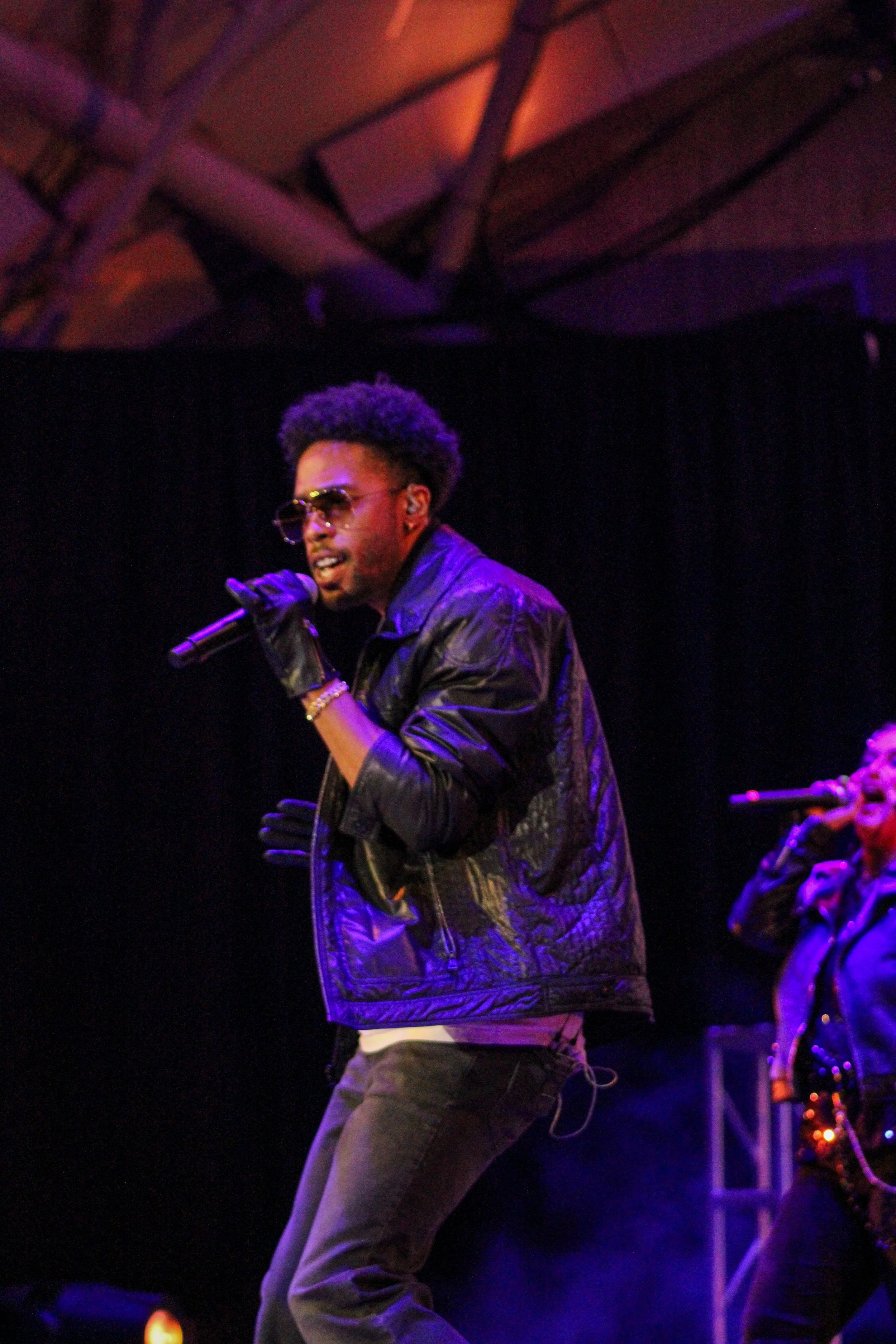 A male performer singing into a microphone on stage, wearing sunglasses, a black leather jacket, and gloves, with a woman singing in the background.