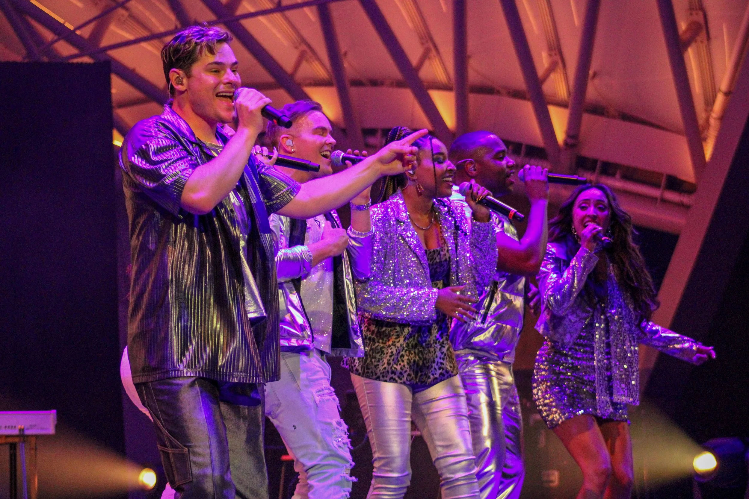Group of musicians singing and performing on stage with microphones, wearing shiny and colorful outfits, under vibrant stage lighting.