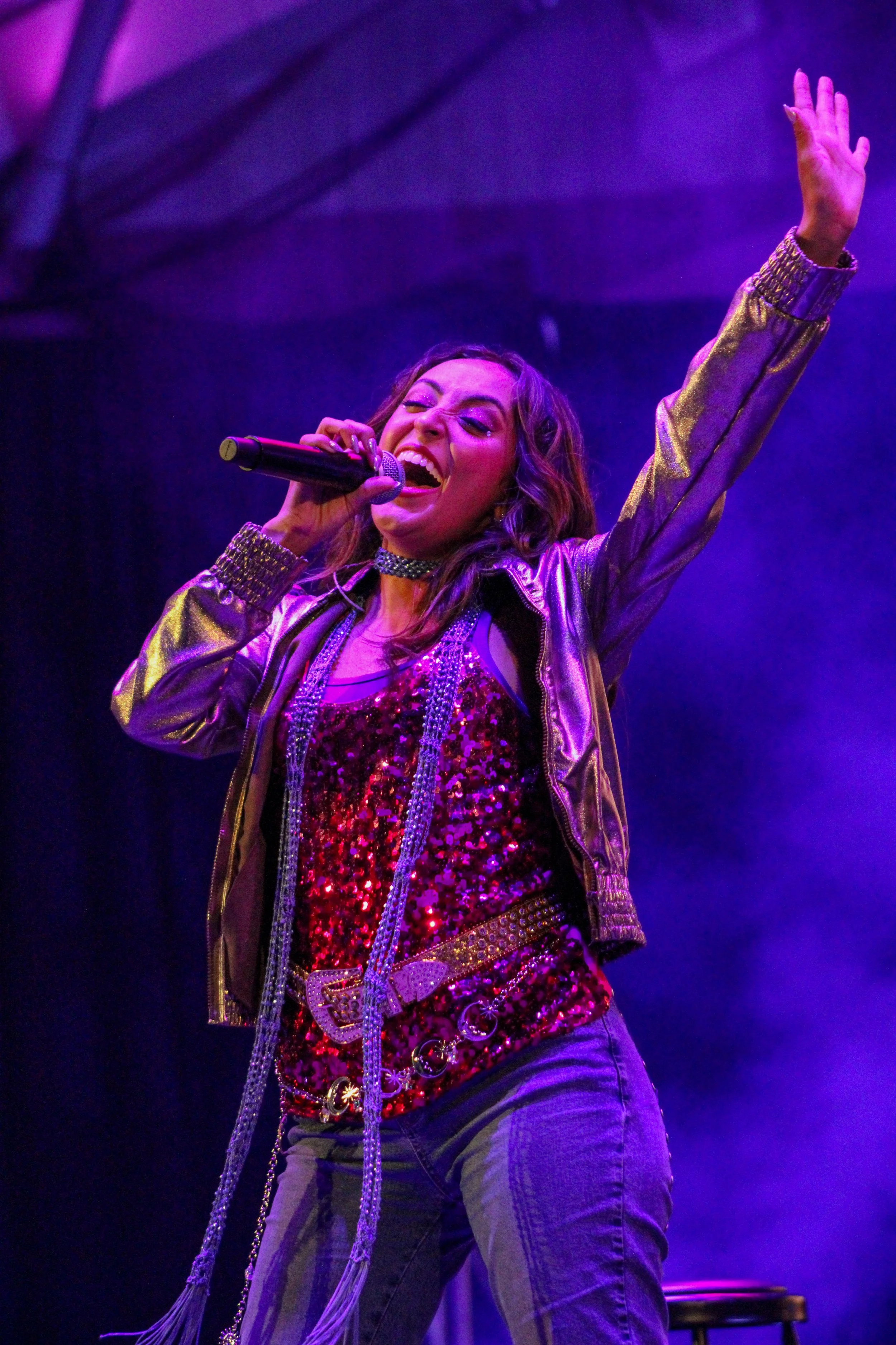 A woman performing on stage, singing into a microphone with her eyes closed and one arm raised, wearing a shiny gold jacket and a red sequined top.