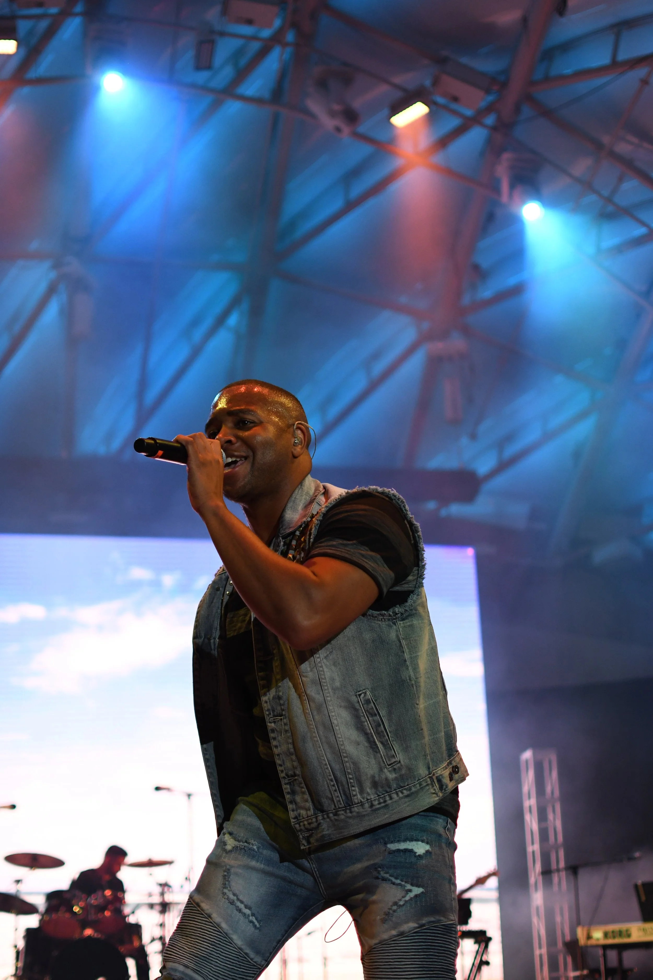 A male singer performing on stage with a microphone, wearing a denim vest and ripped jeans, under blue and purple stage lighting, with a drummer in the background.