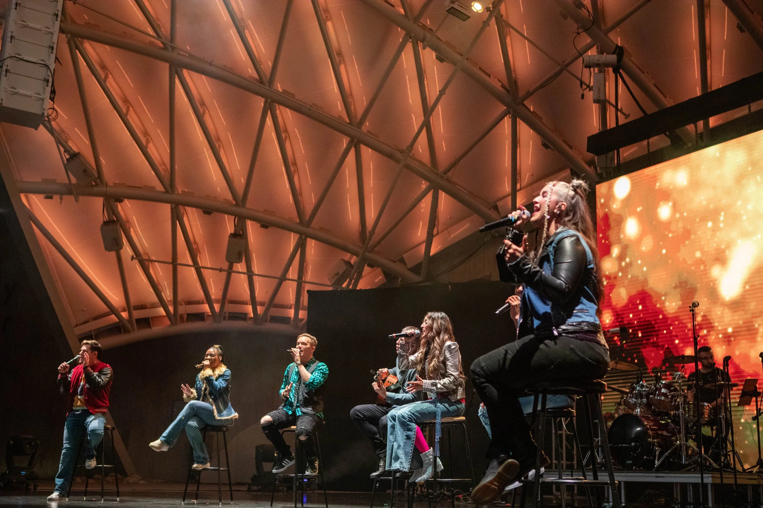 A group of seven singers and one drummer performing on stage with microphones and musical instruments, with a large digital screen in the background displaying orange and yellow abstract visuals, under an arched ceiling.