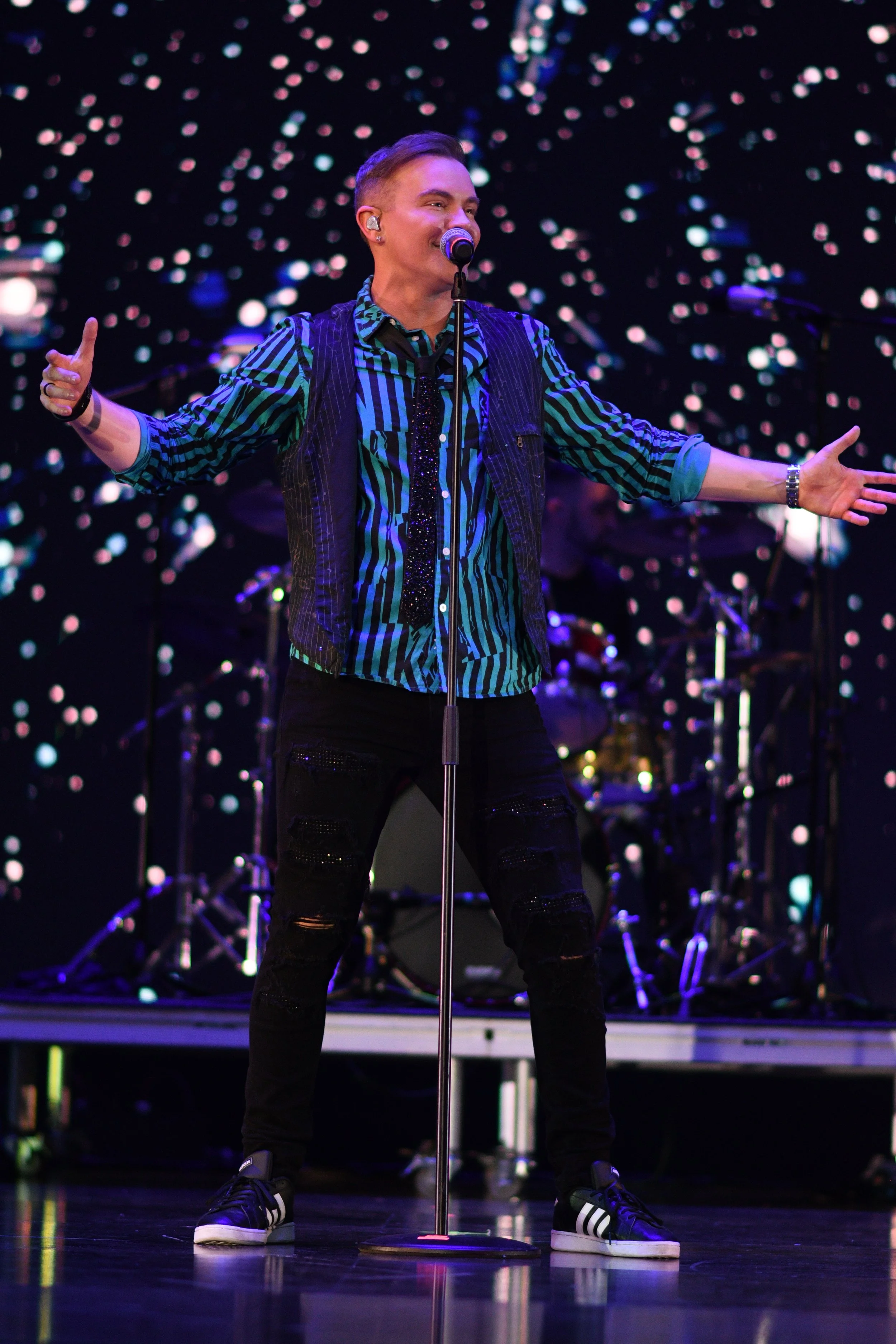 A male performer on stage singing into a microphone, with a colorful, sparkly background and a drummer in the background.